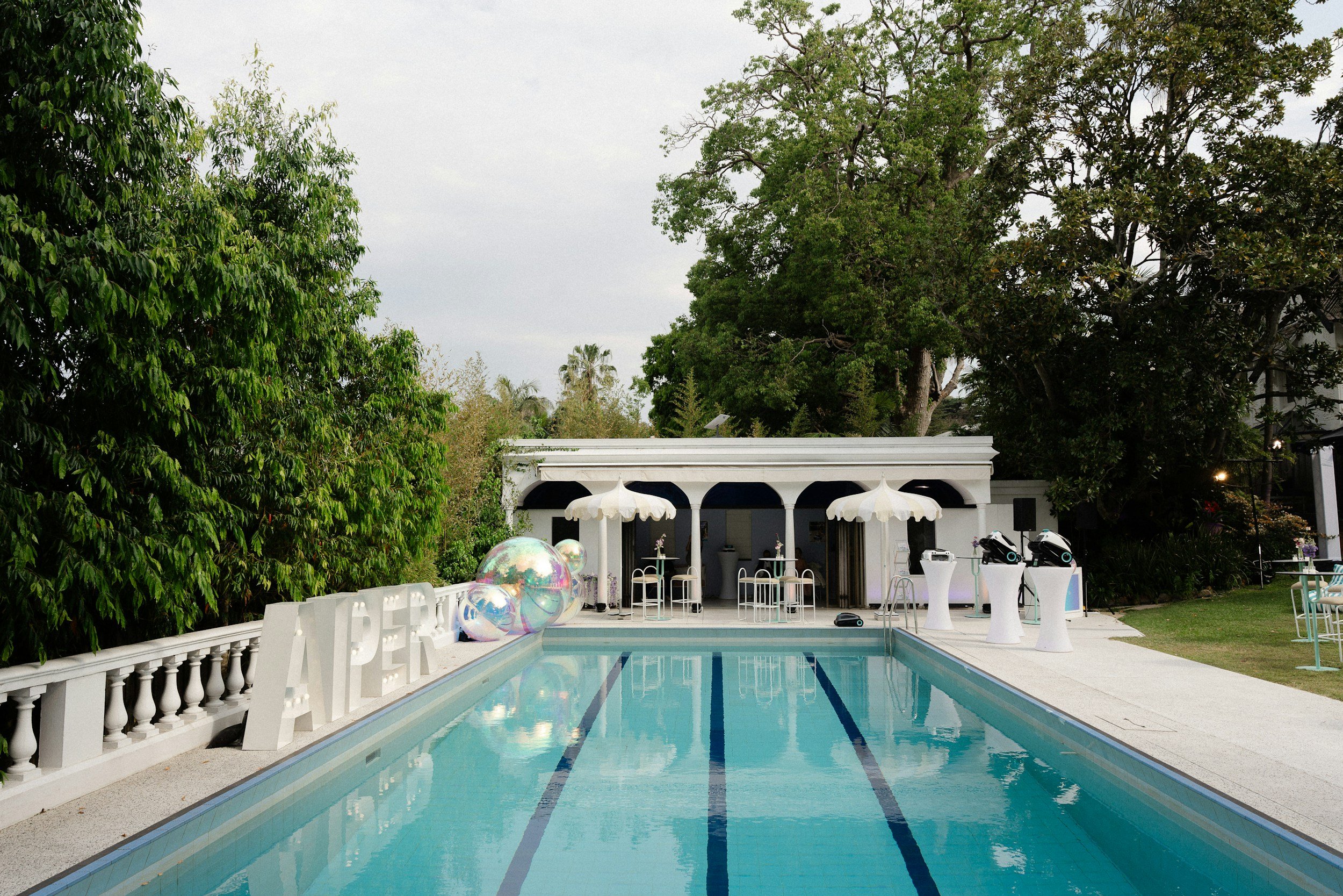 Empty swimming pool with white balustrade, large colorful balloons, and an elegant pool house with white umbrellas and chairs, surrounded by greenery.