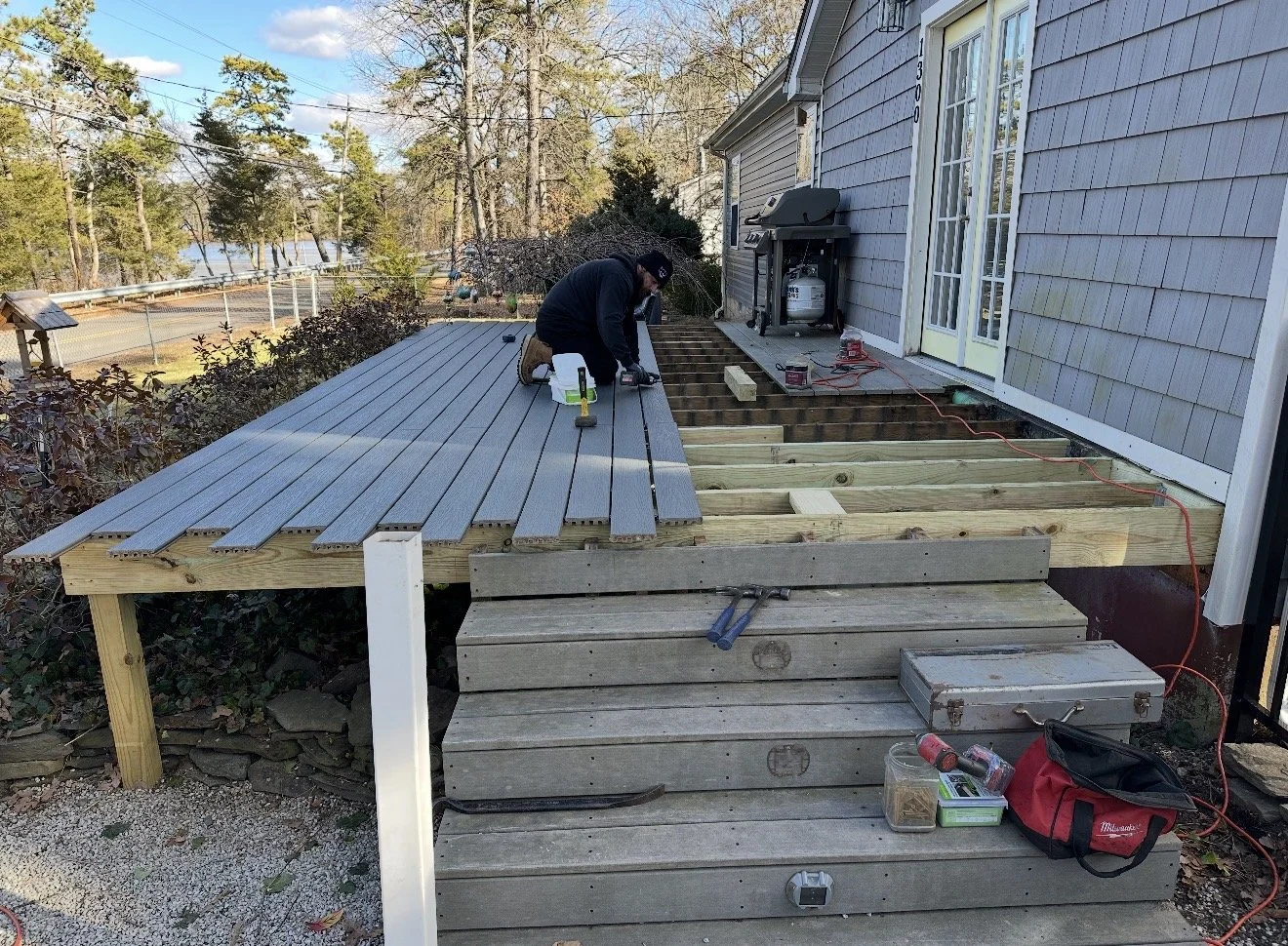 Person installing outdoor decking on a wooden platform attached to the house, with tools and materials around.