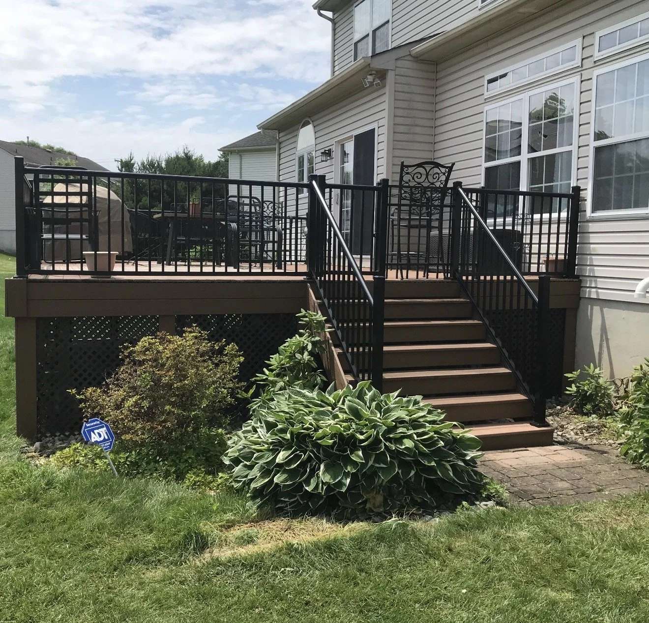 Backyard deck with stairs, black metal railing, outdoor furniture, grill, and surrounding plants.