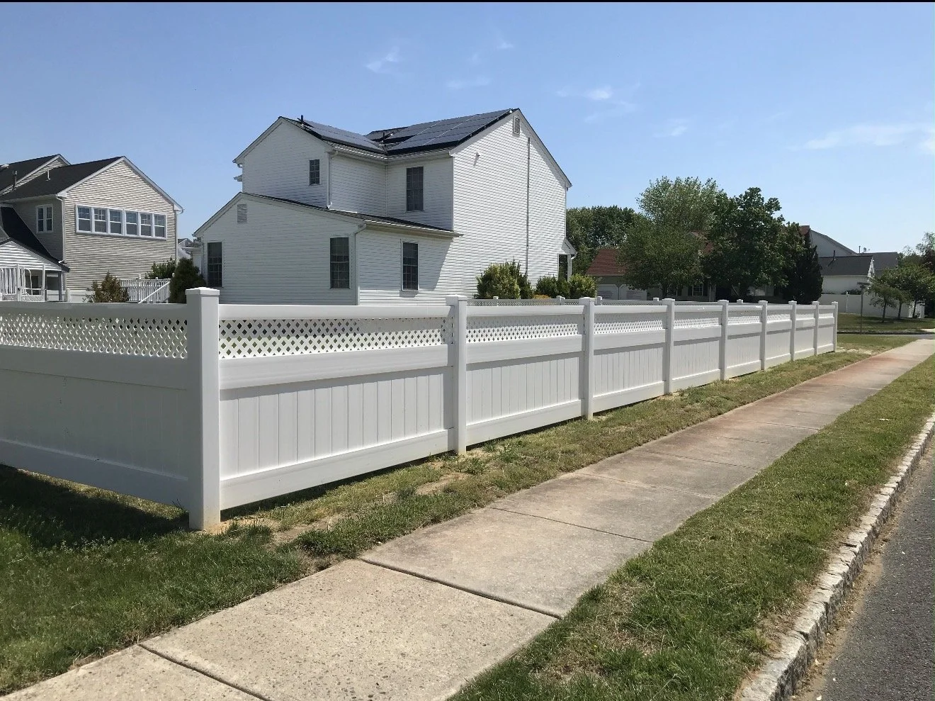 A white wooden fence along a sidewalk in a suburban neighborhood with houses and trees in the background.