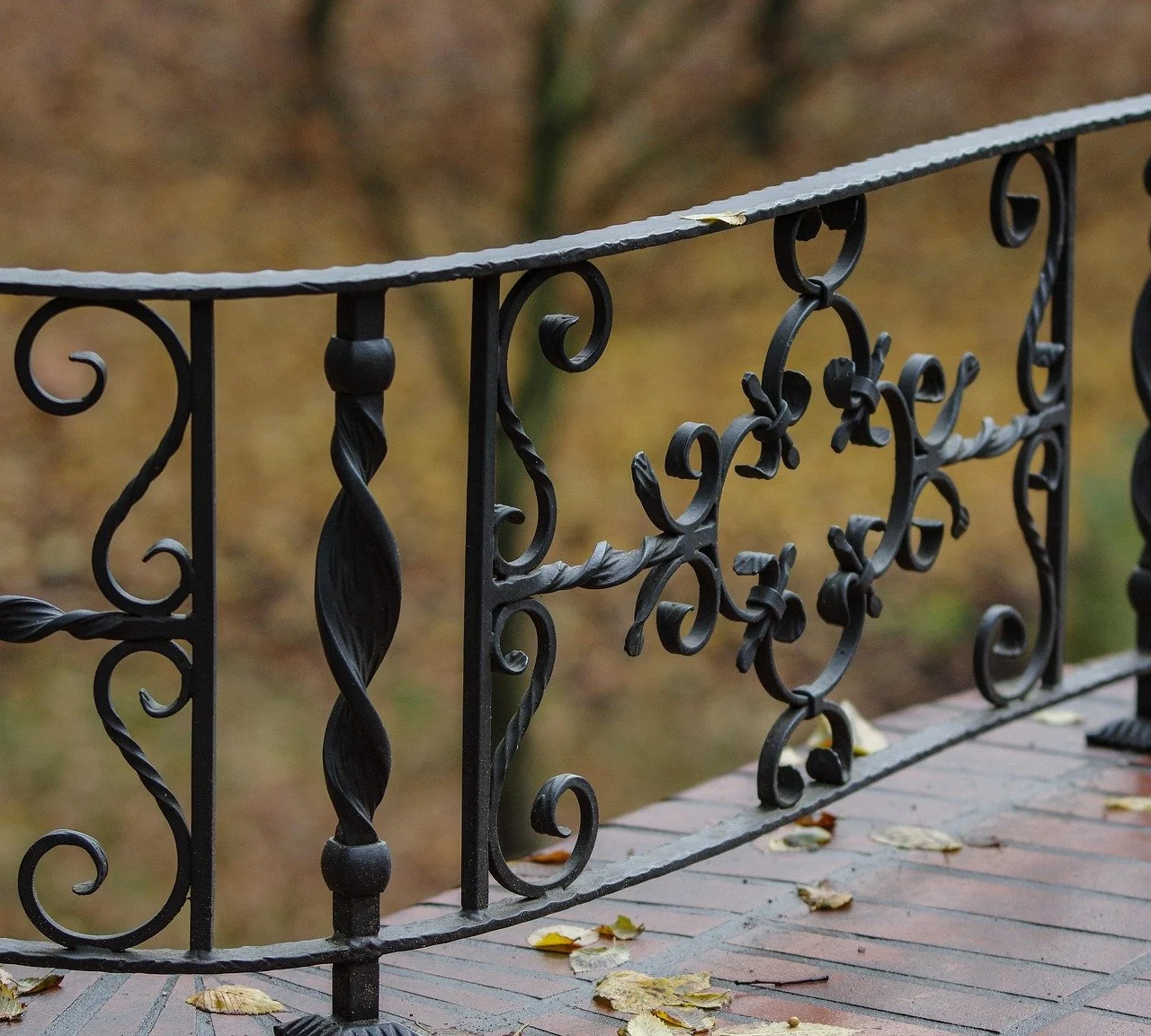 Close-up of a decorative black wrought iron railing on a brick porch with fallen autumn leaves, with blurred trees in the background.