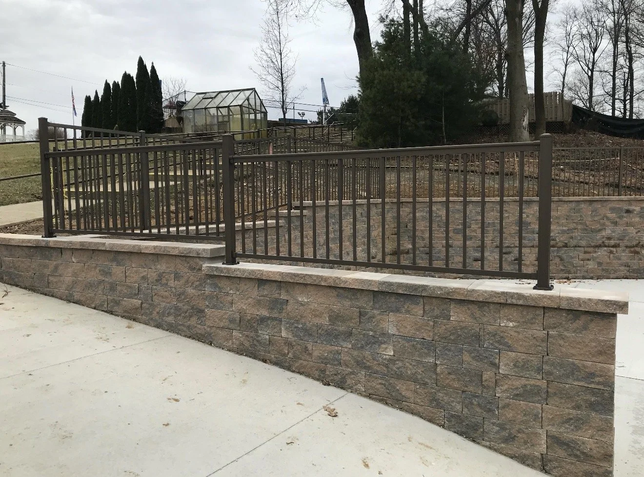 A sidewalk adjacent to a stone retaining wall with metal fencing on top, overlooking a garden area with trees and a greenhouse in the background.