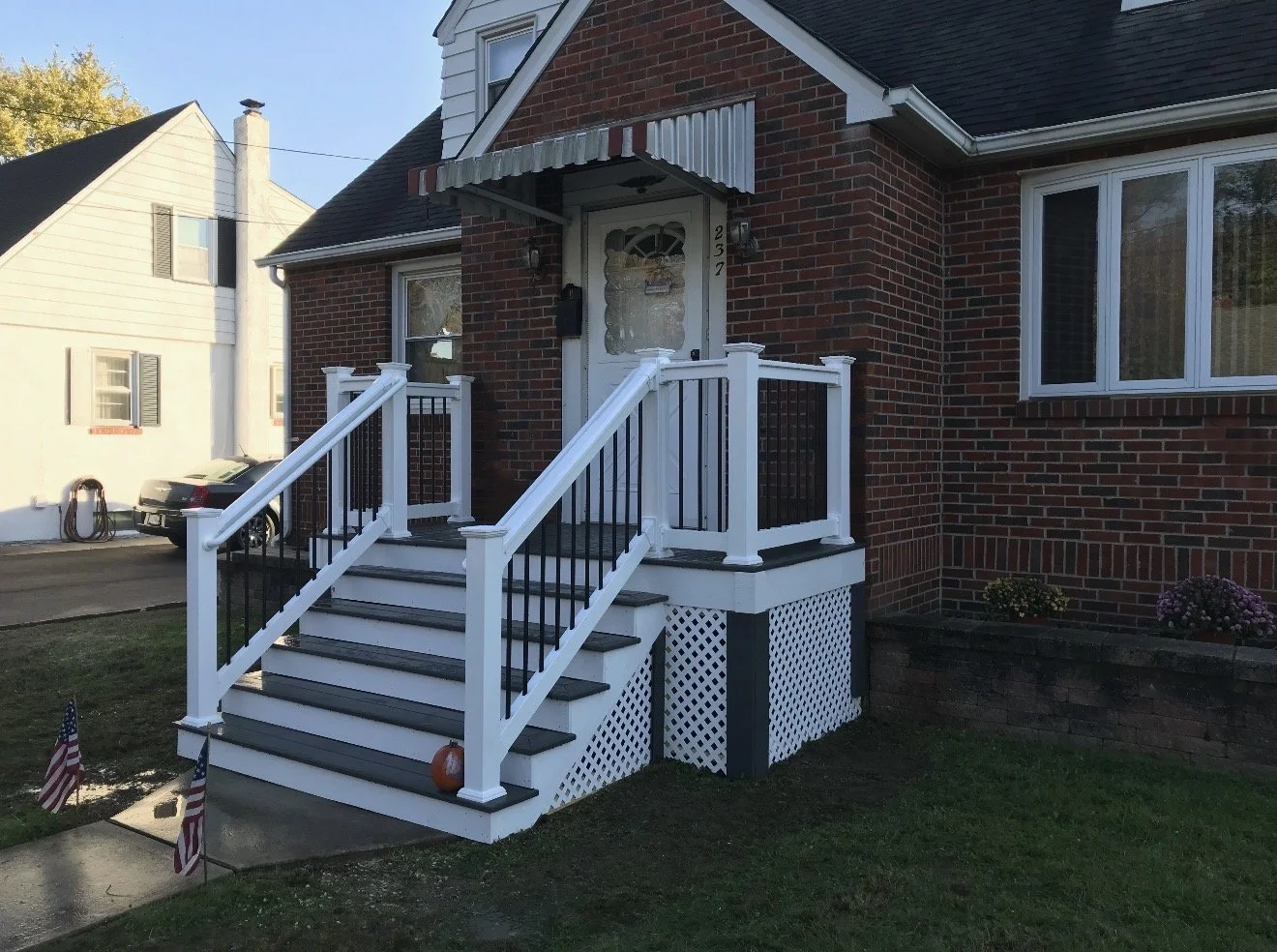 Brick house with white staircase and porch, black and white railing, and striped awning above the door, residential neighborhood with neighboring houses, American flags, and autumn trees.