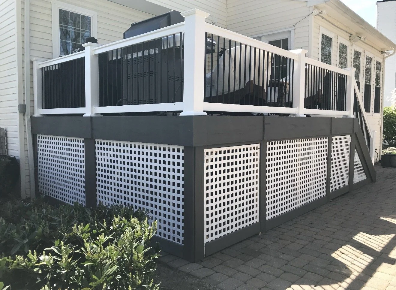 Exterior view of a house with a backyard porch, featuring a raised deck with black and white railing, a staircase leading up to the deck, and lattice skirting below. Paved patio stones extend around the deck, with some greenery in the foreground.