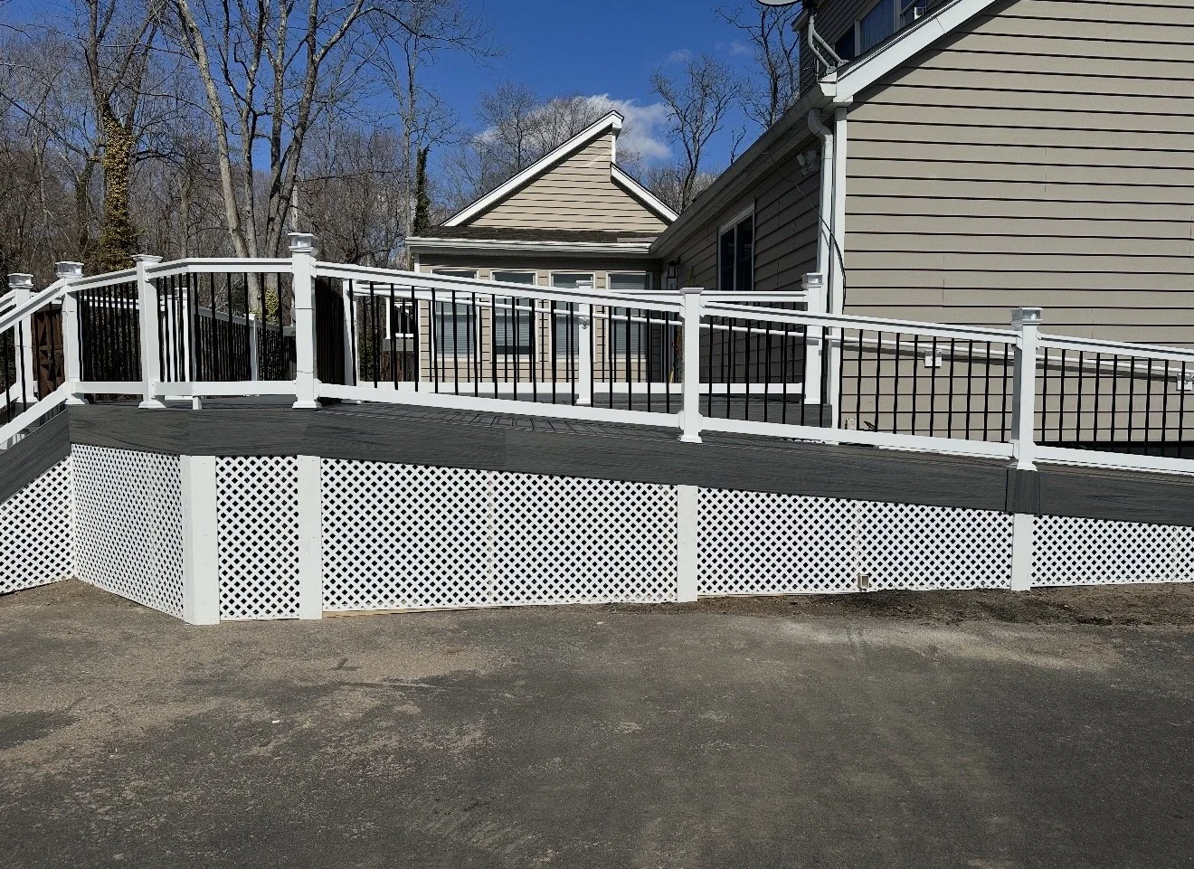 A residential backyard with a raised wooden deck that has white and black railings, stairs, and lattice skirting, surrounded by a house with beige siding, leafless trees and a bright blue sky in the background.