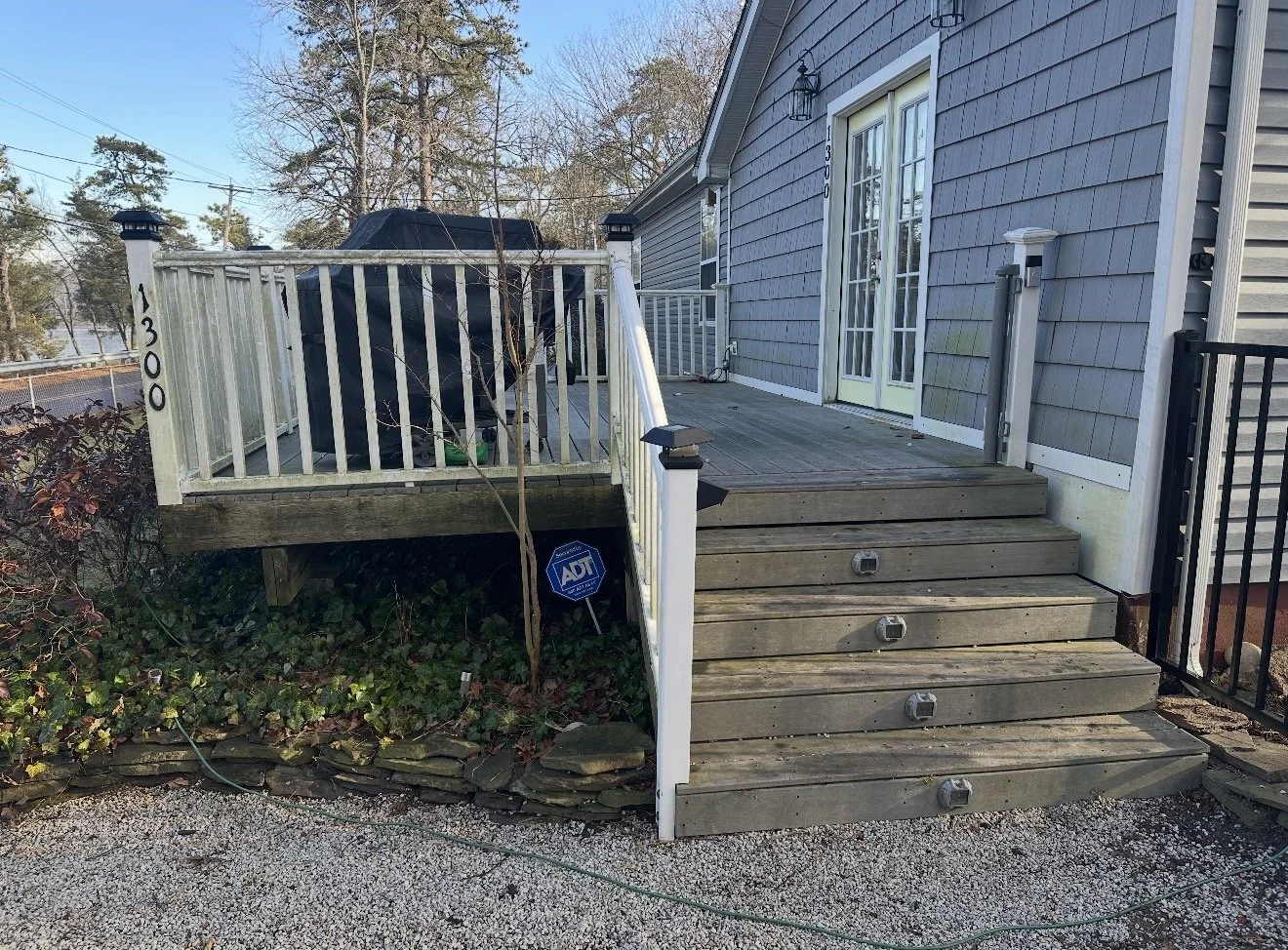 Exterior view of a house with a wooden deck and stairs leading to a glass double door, surrounded by a garden with a path, an ADT security sign, and a blue-gray siding house with black and white outdoor lighting fixtures.