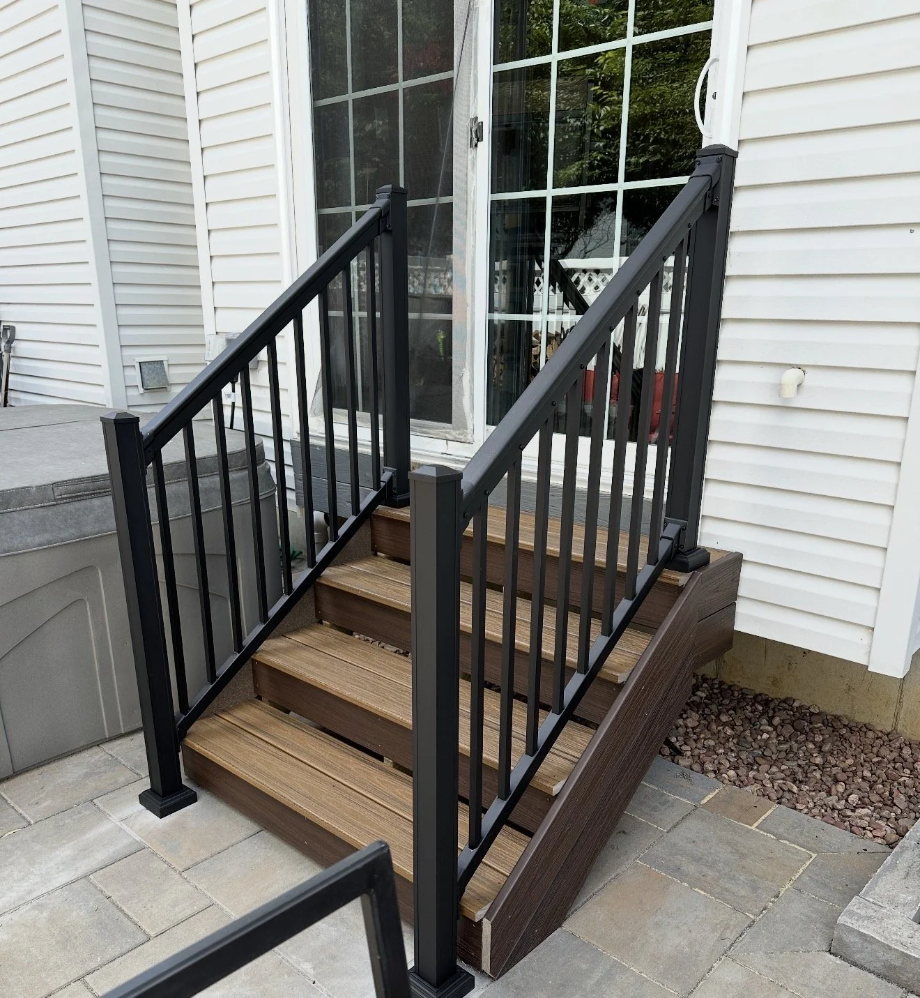 Wooden stairs with metal black railings leading up to a glass sliding door on a house with white vinyl siding.