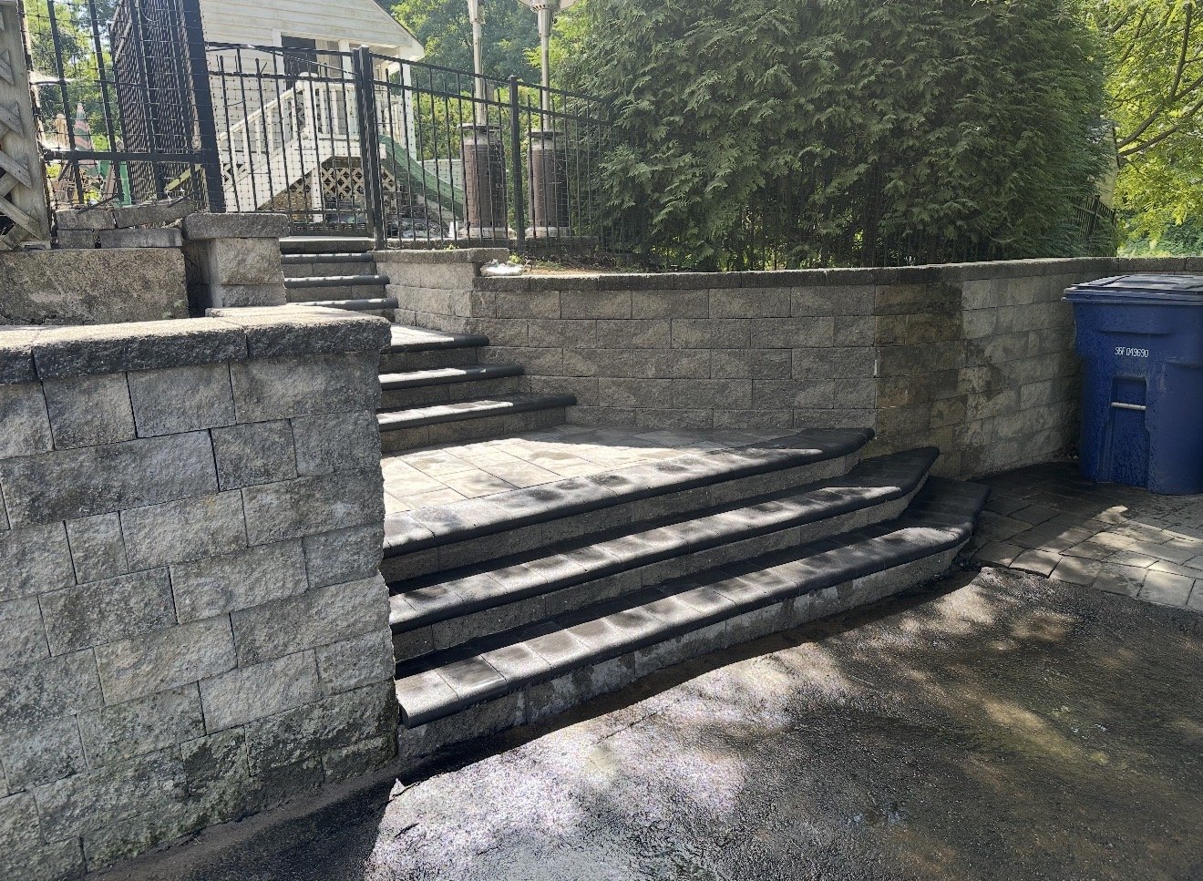 Stone steps leading up to a higher level, with a black metal fence and lush greenery in the background, and a blue recycling bin on the side.