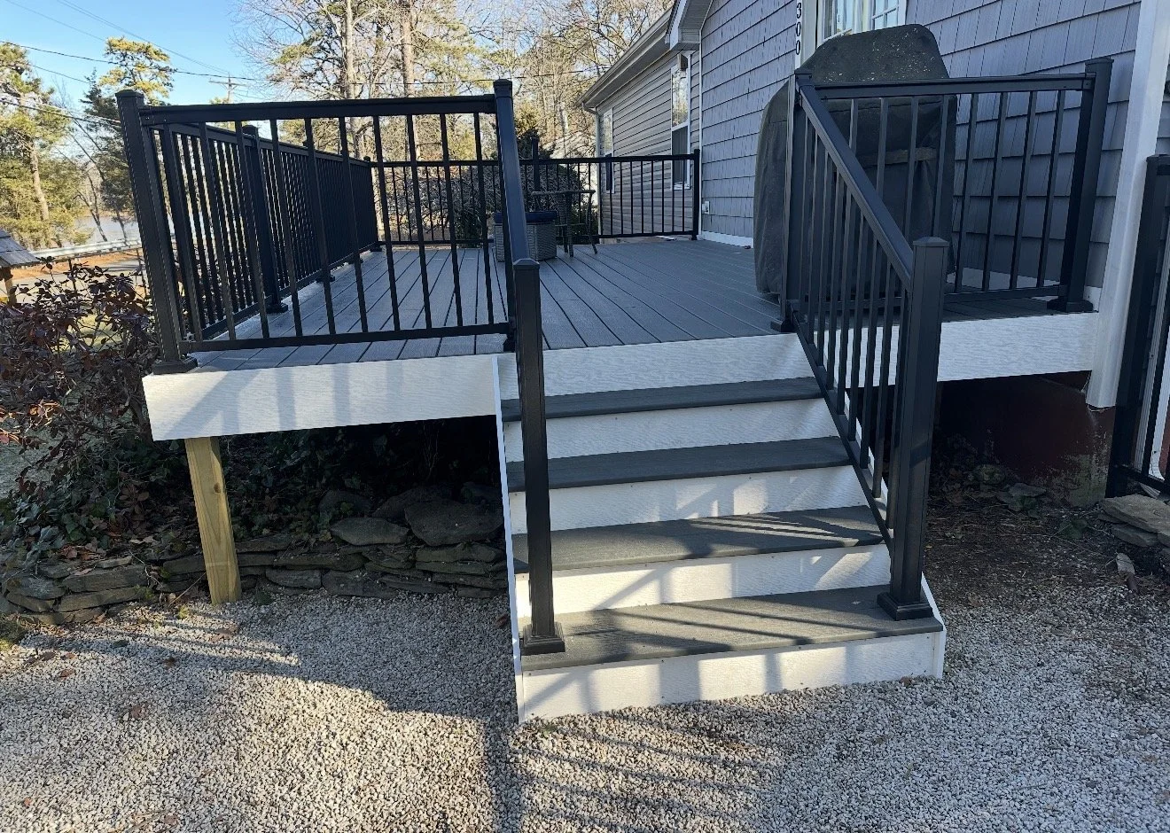 Wooden deck with black metal railing, a set of steps leading down to gravel, attached to a gray house with horizontal siding, and a gas grill covered with a black cover on the deck.