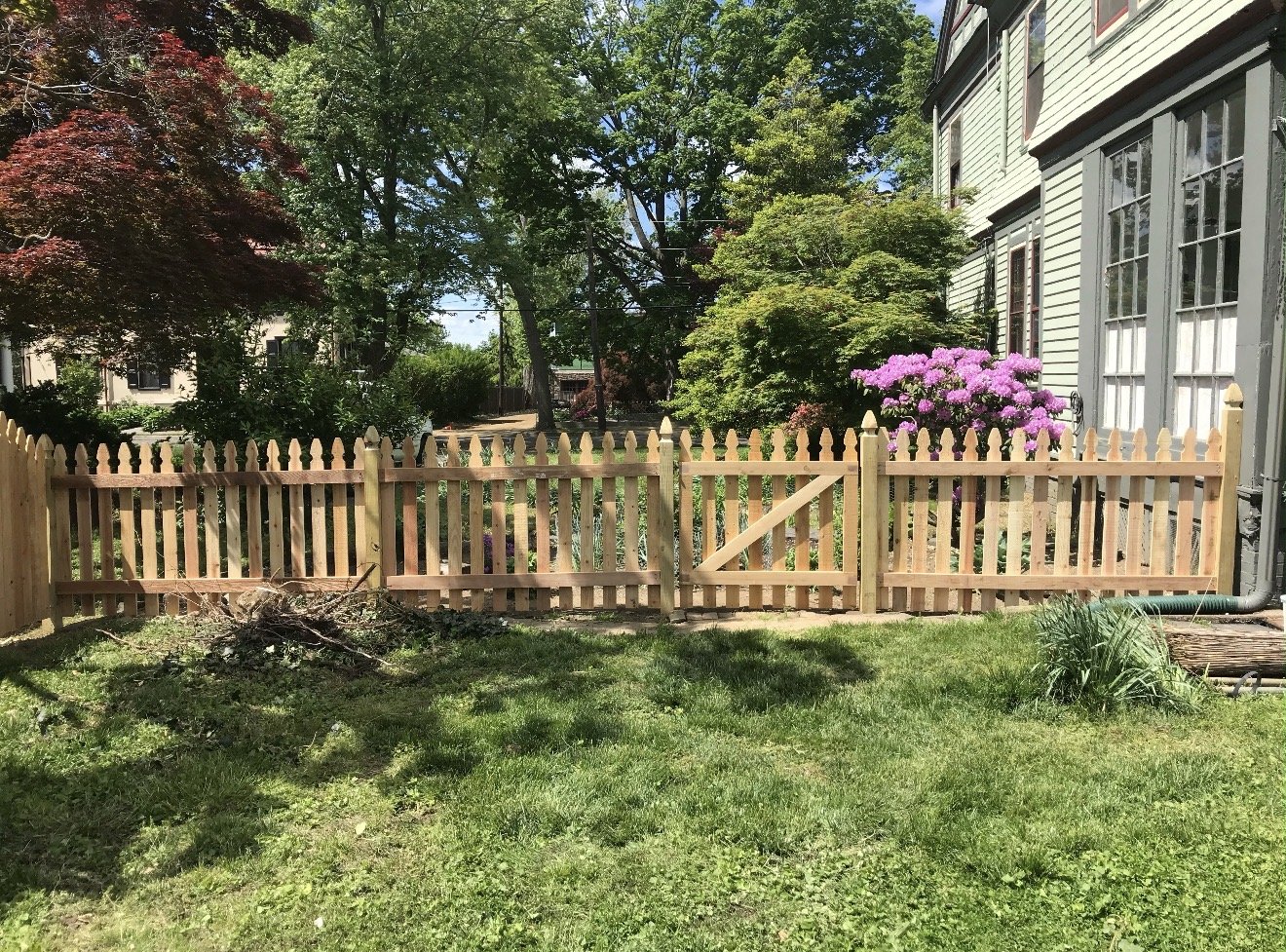A backyard scene with a curved brick pathway, black metal fence, green lawn, trees, a house, and a sunset sky with a sun halo