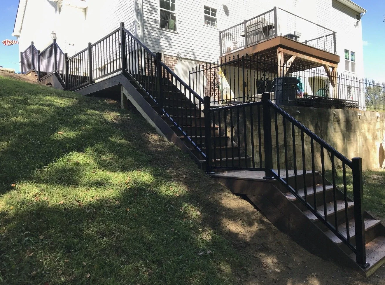 Exterior view of a house with a staircase and balcony. The staircase has black railings and leads up a grassy slope to a back porch area. The house is white with multiple windows.
