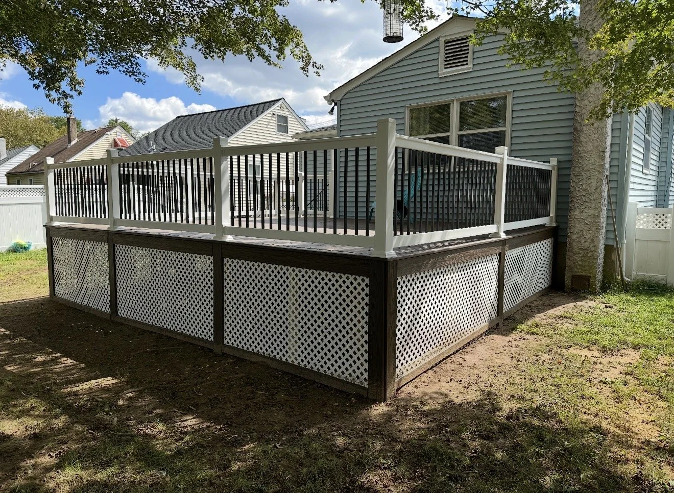 A backyard deck with white fencing and lattice skirting, attached to a blue house, with trees and neighboring houses visible in the background.