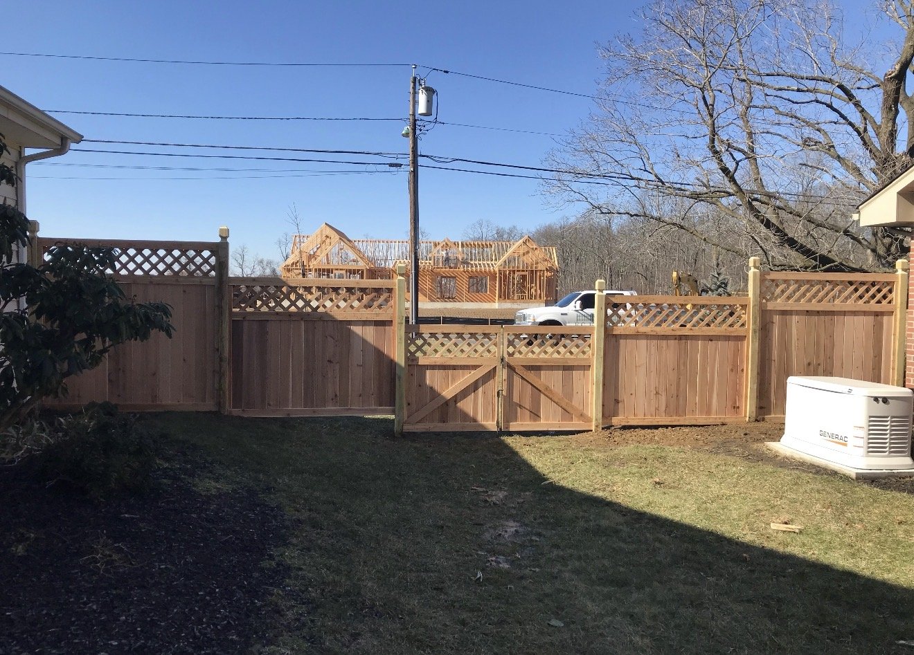 View of a backyard enclosed by a wooden fence. Behind the fence, there is a construction site with a wooden framework for a building. A white truck is parked beyond the fence. The sky is clear and blue, and there are tall, leafless trees in the backg