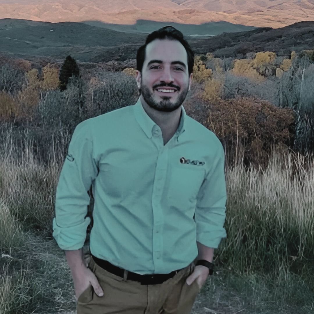 Un hombre con camisa de manga larga y cinturón, sonriendo en un paisaje de montañas con árboles y hierba seca, en un atardecer.