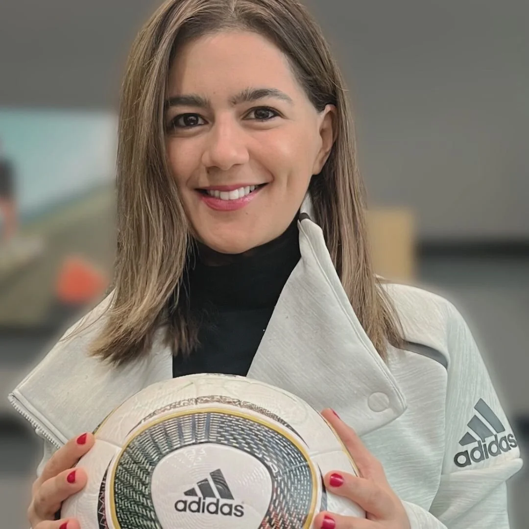 Mujer sonriendo, sosteniendo un balón de fútbol con el logotipo de Adidas, lleva chaqueta blanca y fondo difuso.