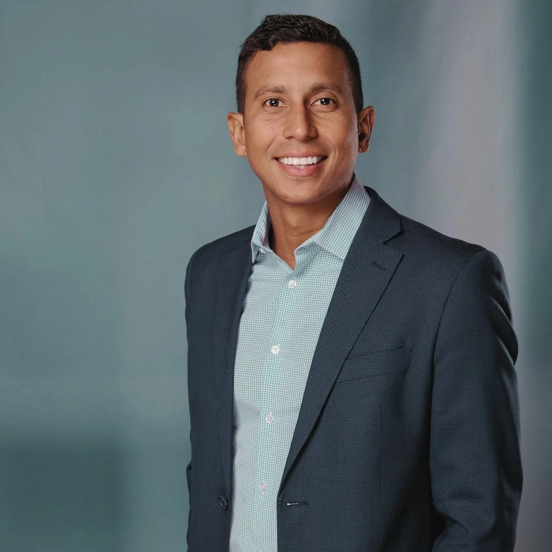 Hombre sonriente con traje y camisa, fondo de color neutro.