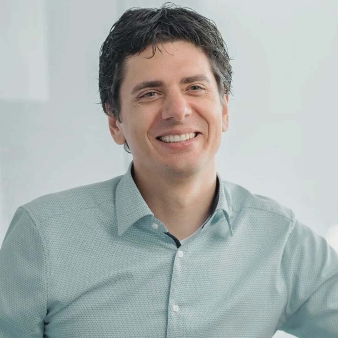 Hombre con cabello castaño oscuro, sonrisa, camisa de vestir azul claro, fondo blanco.