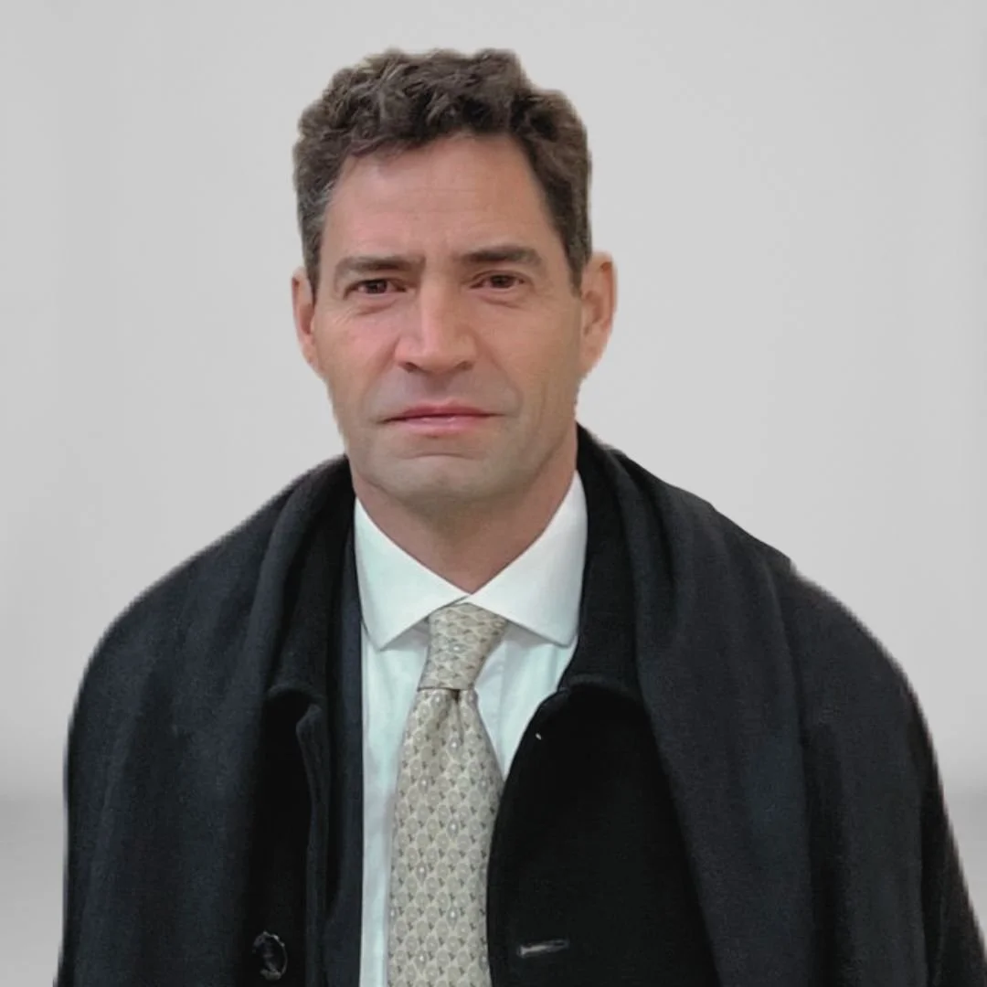 Hombre con cabello oscuro y expresión seria, vestido con camisa blanca, corbata y abrigo negro, de fondo gris.