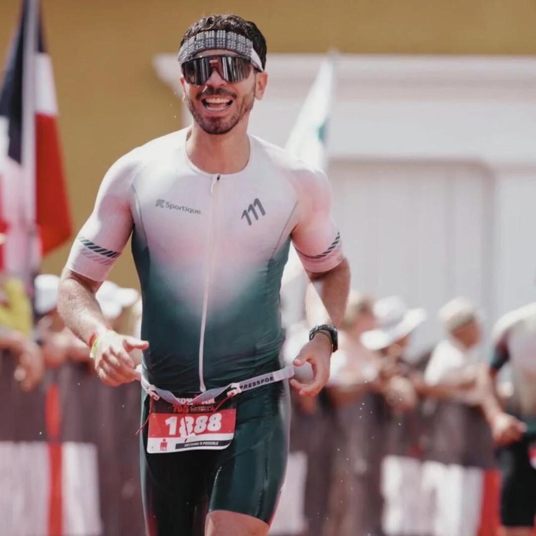 Hombre sonriendo y corriendo en una competencia, lleva un traje deportivo, gafas oscuras y una cinta en la cabeza, con un número de corredor en el frente.
