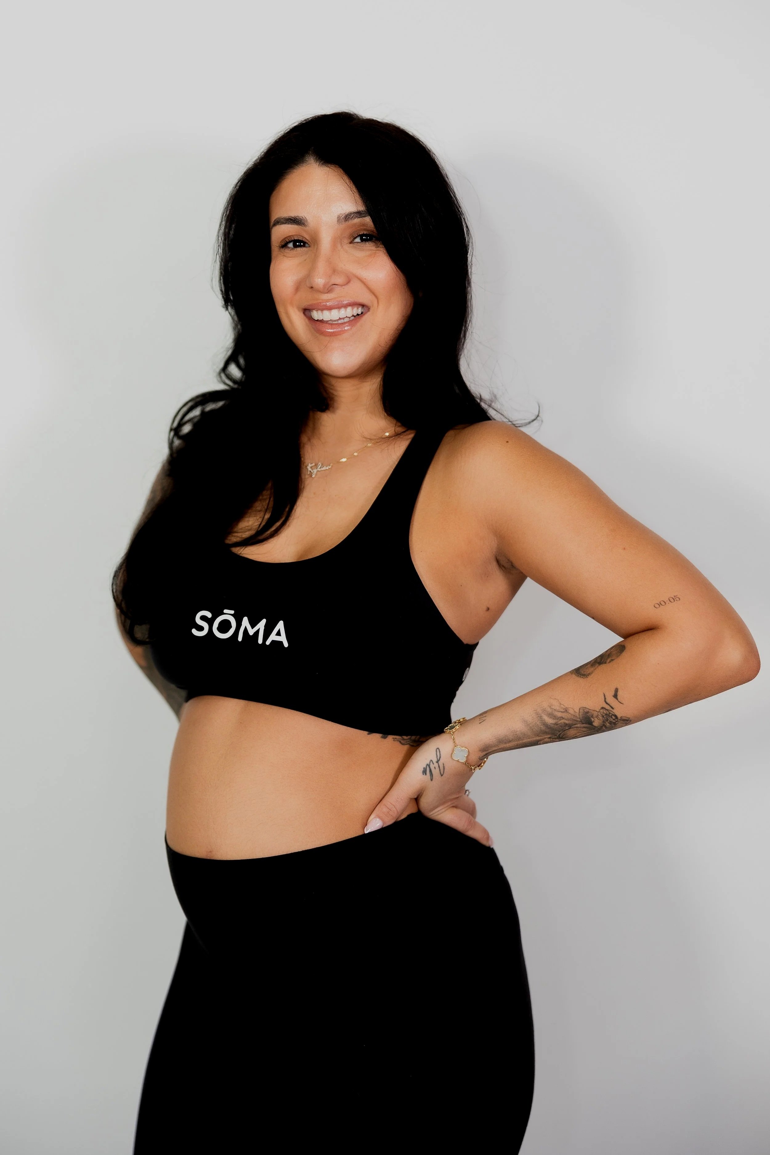 A woman with long black hair smiling, wearing a black sports bra with the word 'SÖMA' and black leggings, standing against a plain light grey background, with tattoos on her left arm and a gold bracelet on her right wrist.