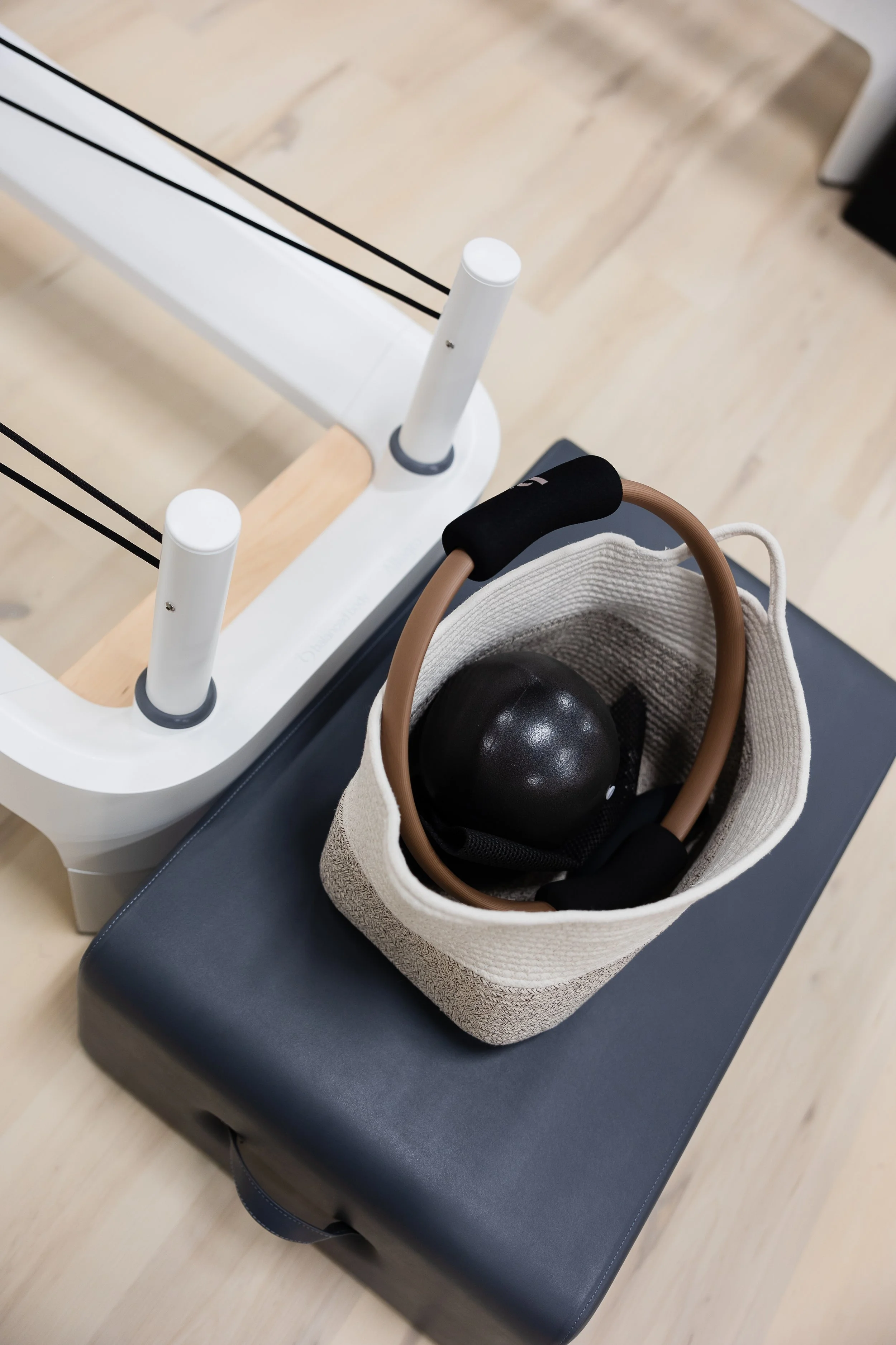A white pilates reformer on a light wood floor with a beige tote bag containing a brown handle and black spherical objects on a black padded bench.