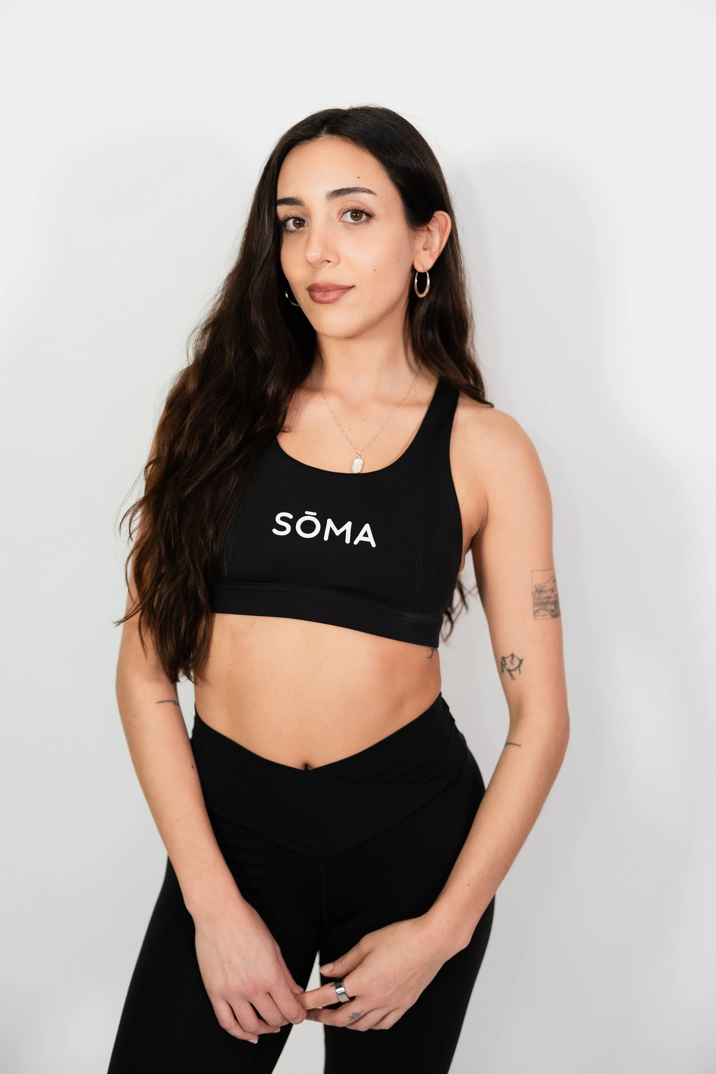 A young woman with long wavy brown hair, wearing a black sports bra with the word 'SÖMA' on it, and black high-waisted pants, standing against a plain white background. She has hoop earrings, a necklace, and tattoos on her left arm.