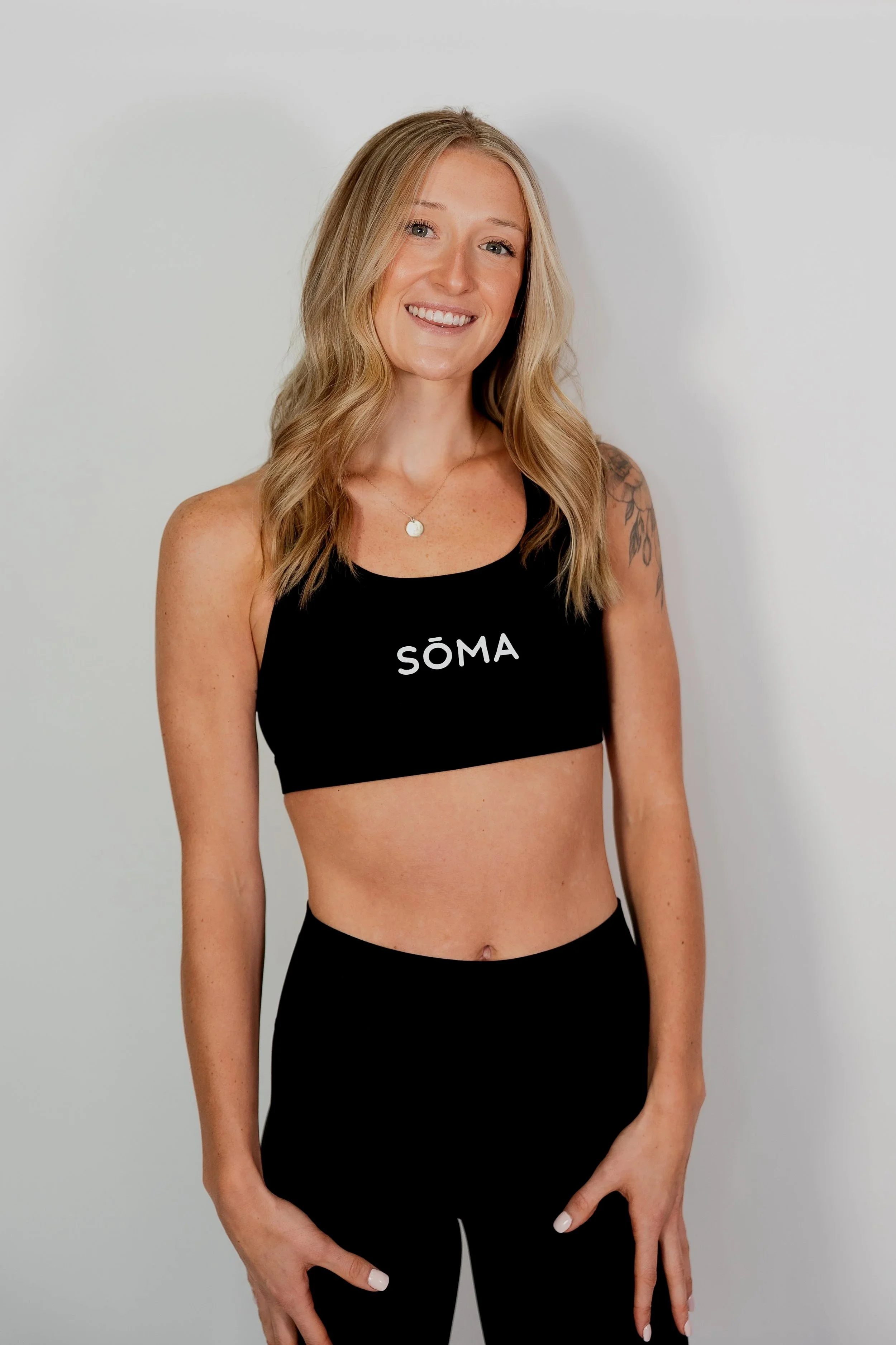 A woman with long blonde hair, blue eyes, and a wide smile, wearing a black sports bra with the word 'SÖMA' on it and black leggings, standing against a plain white background.