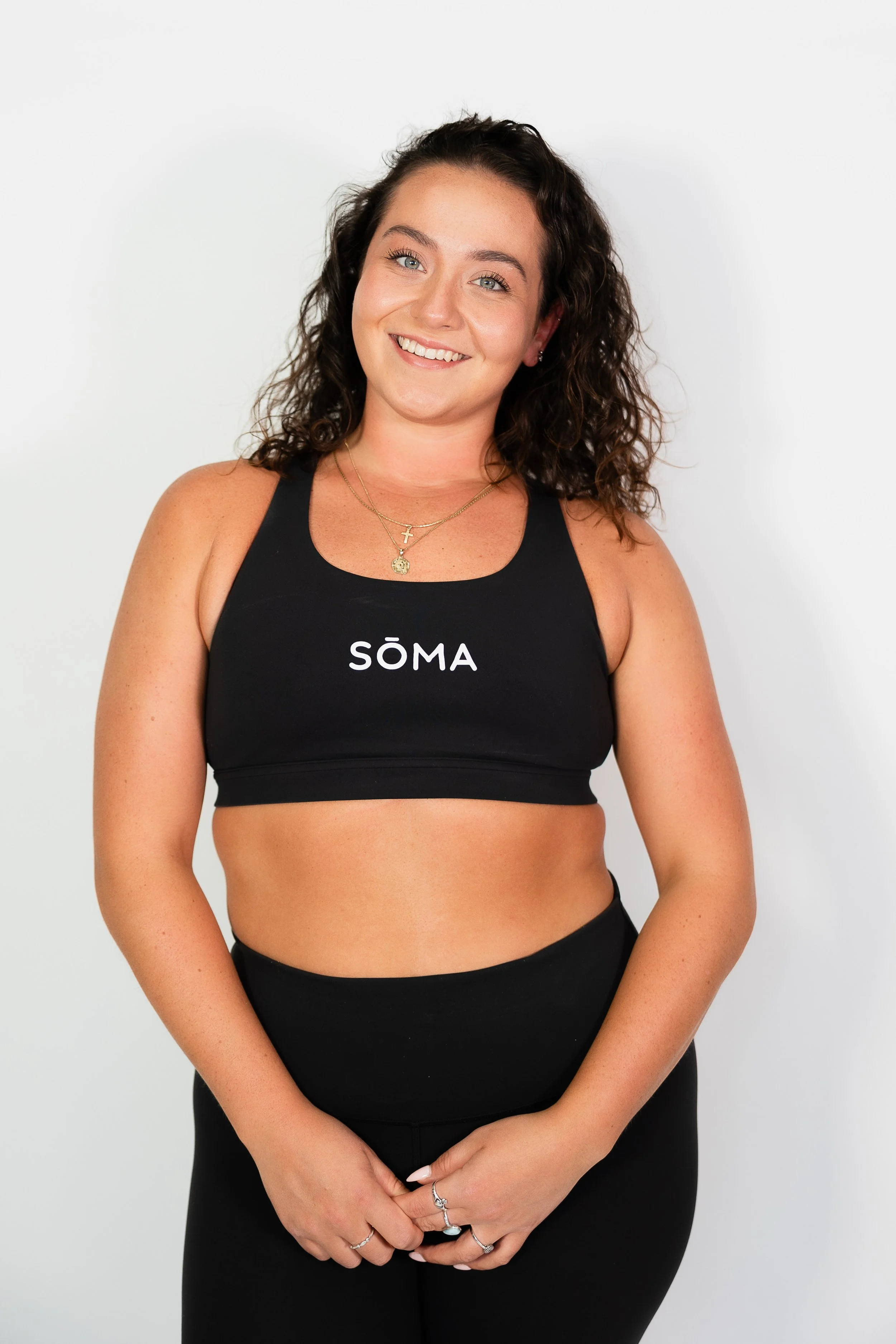 A young woman with curly brown hair and light skin, smiling, wearing a black sports bra with 'SOMA' written on it and matching black leggings, stands against a plain white background.