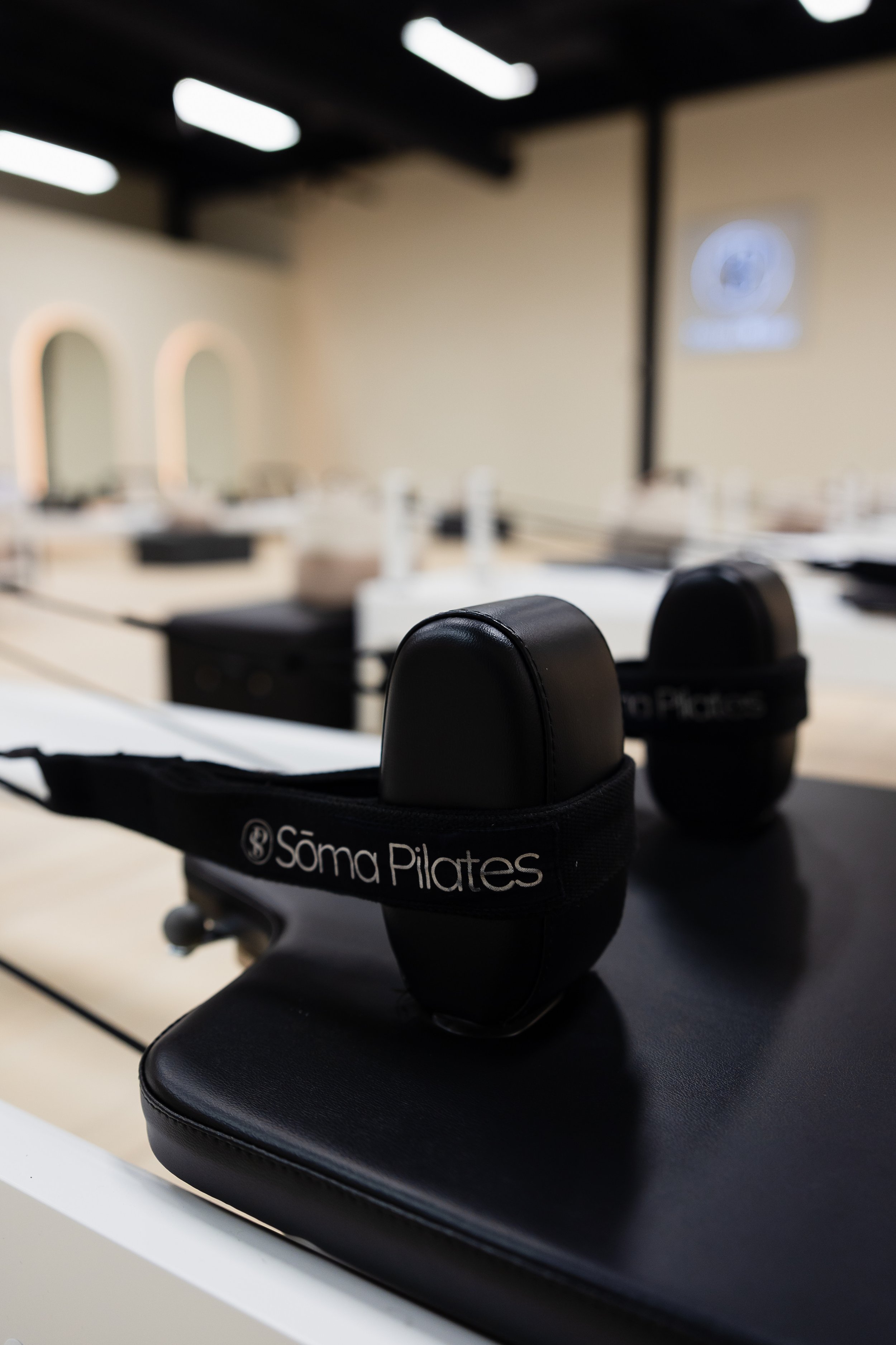 Black suspension trainer with 'Soma Pilates' logo on the strap, placed on a black chair in a Pilates studio, blurred beige wall with circular mirrors and a logo in the background.