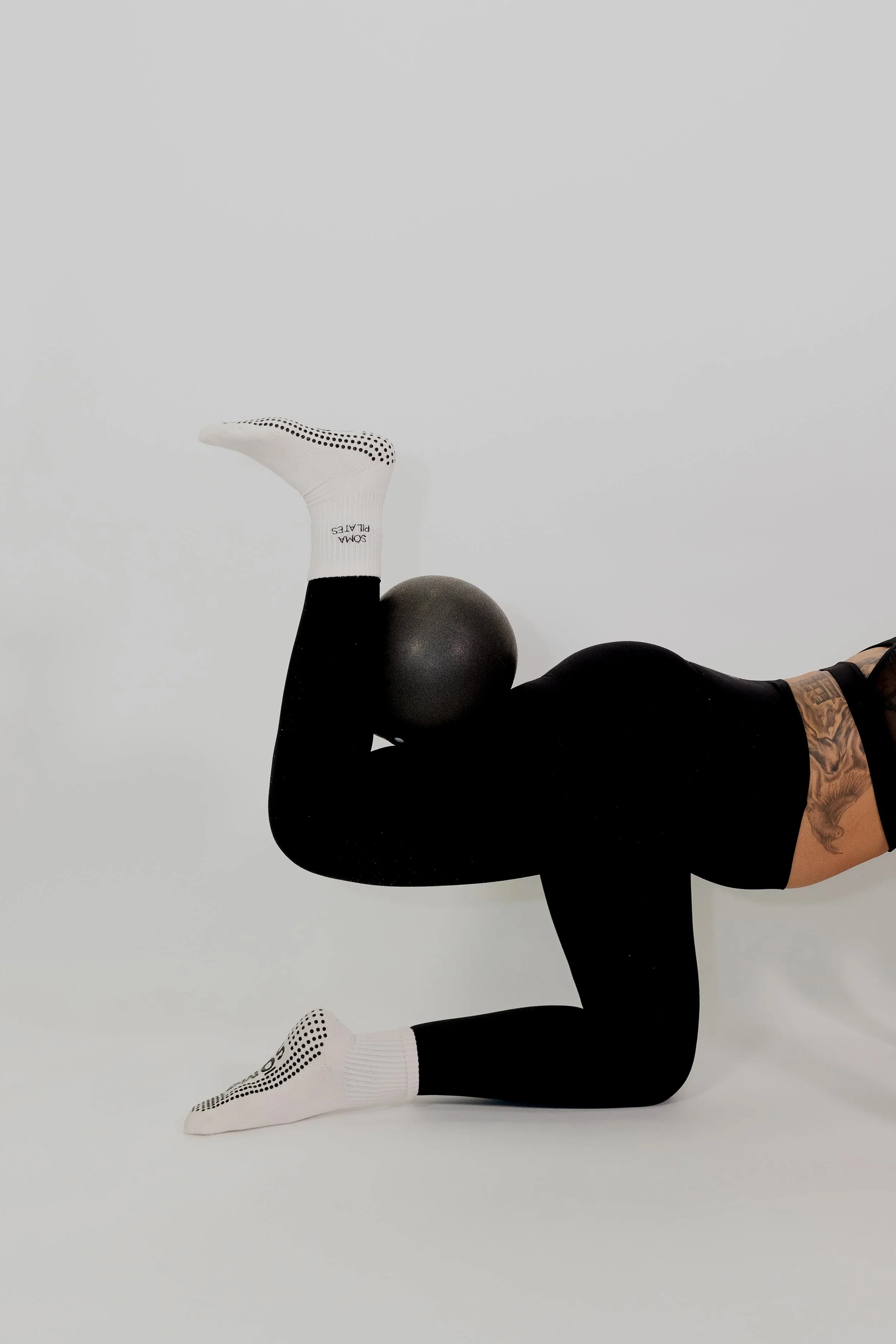 Person lying on the floor doing pilates with legs raised and one arm supporting the body, wearing white socks and black leggings, against a plain white background.