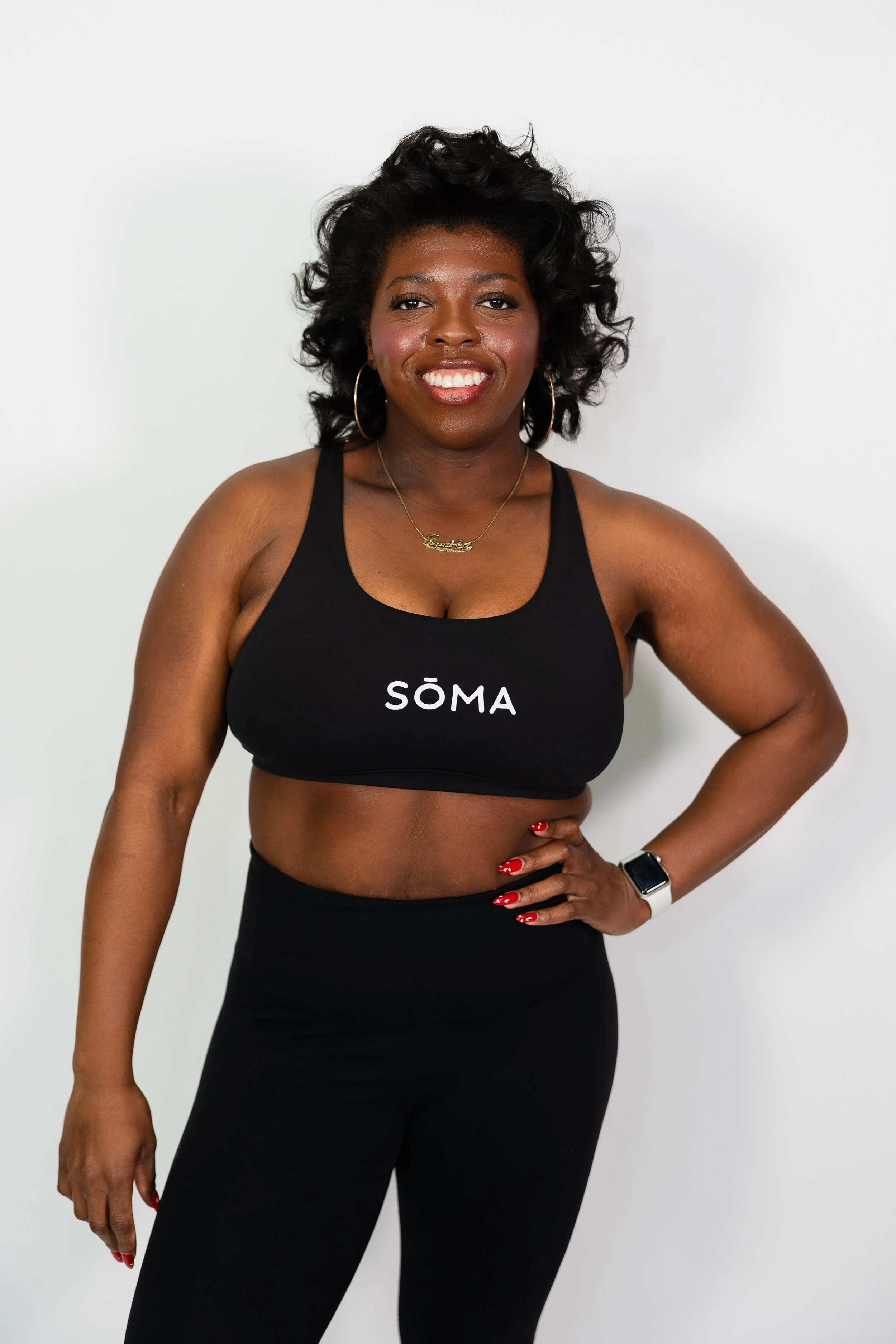 A smiling Black woman with short curly black hair, wearing hoop earrings, a gold necklace, a black sports bra with the word 'SÖMA,' and black pants, posing against a plain white background.