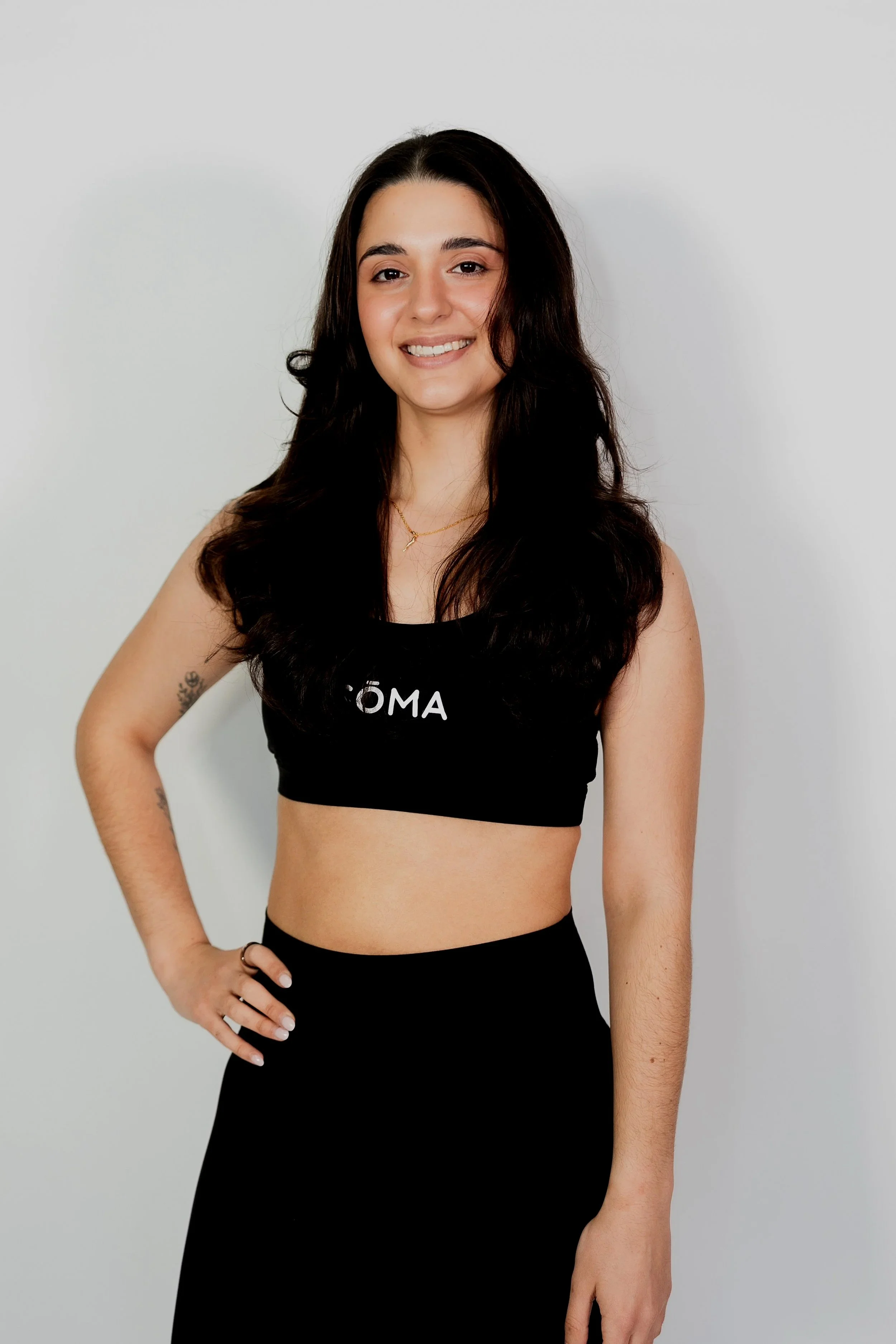 A young woman with long dark hair wearing a black sports bra and a long black skirt, smiling and posing against a plain white wall.