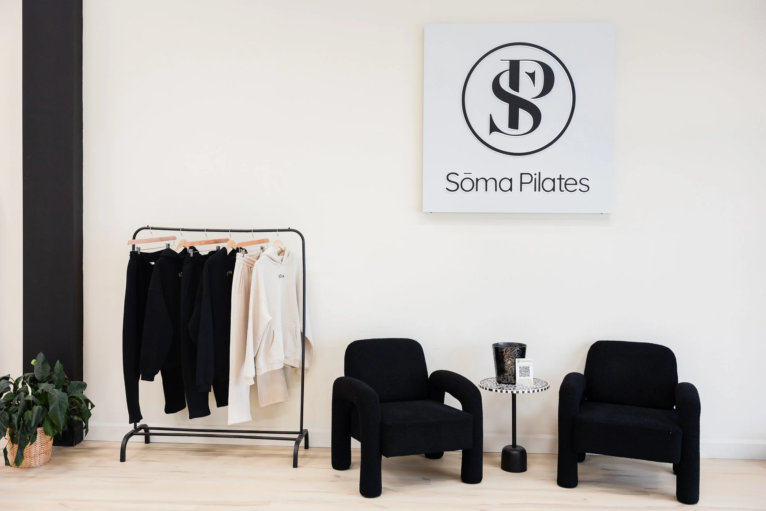 Interior of a Pilates studio named Soma Pilates, featuring a clothing rack with hoodies and sweatpants, a seating area with black chairs and a small round table, and a sign with the studio's logo and name on the wall.