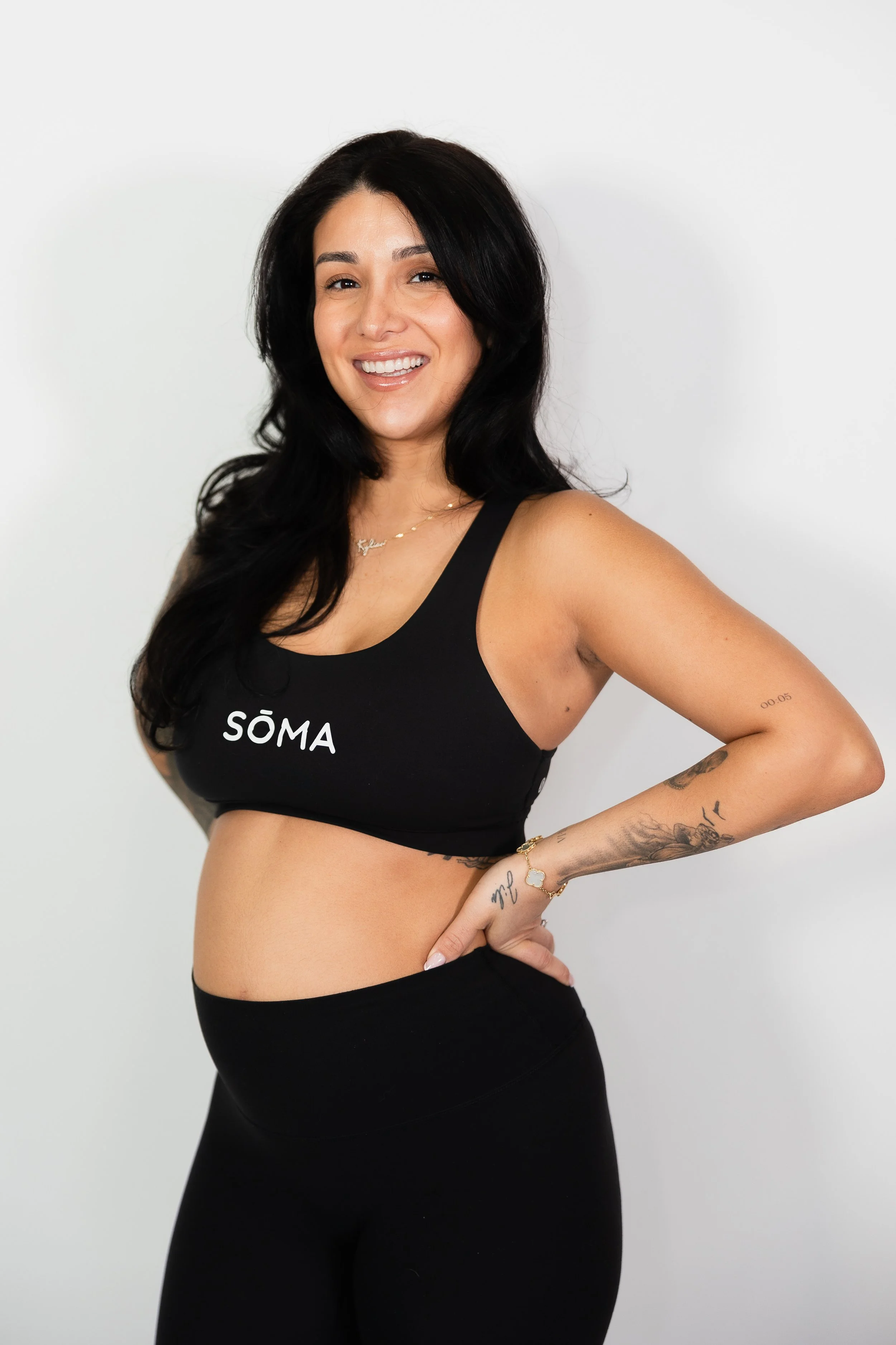 Woman with long black hair wearing a black sports bra and matching leggings, smiling and posing against a plain white background.