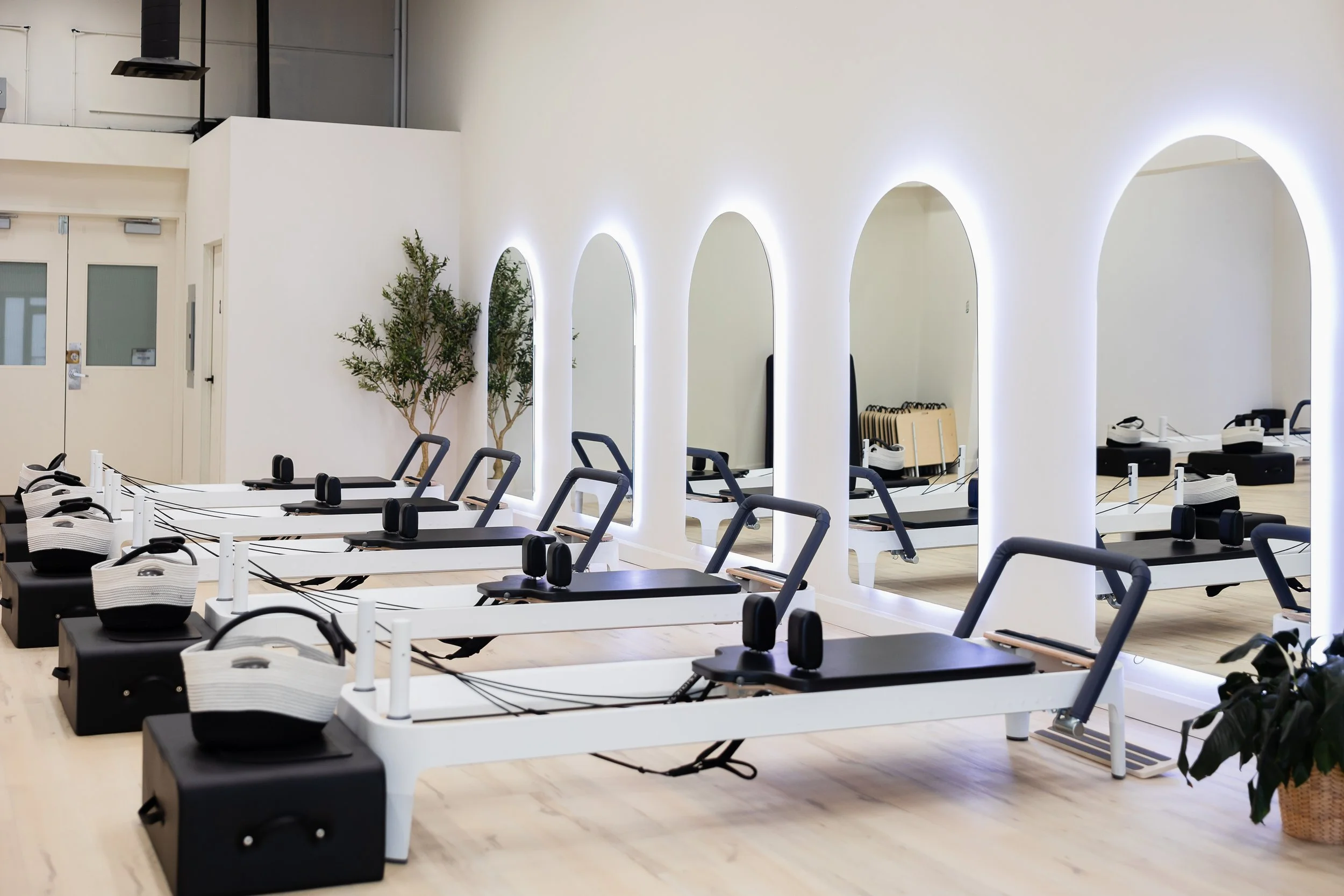 Workout studio with pilates reformer machines arranged in rows, white walls with oval mirrors, potted plants, and bright lighting.