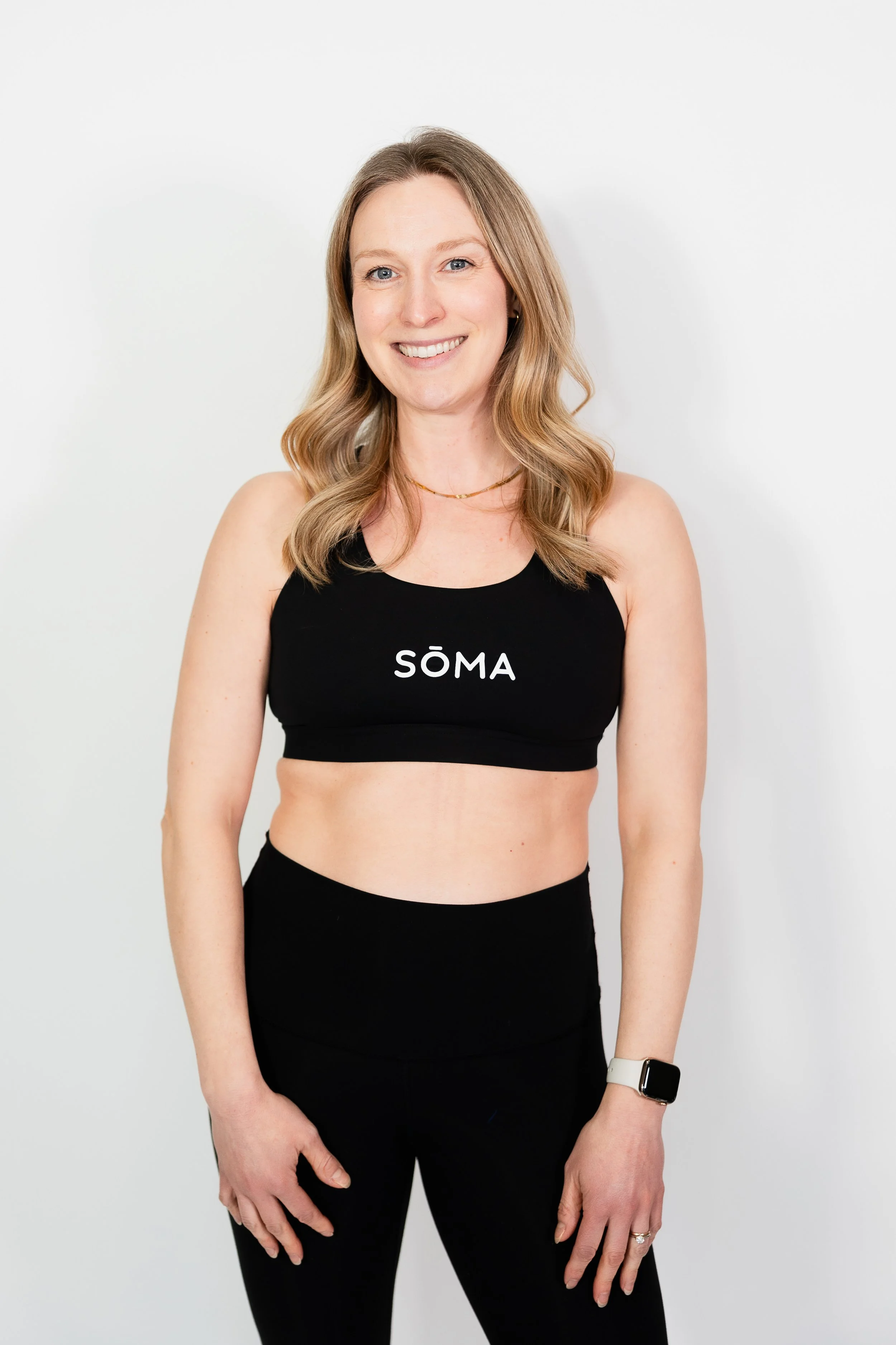 A woman with shoulder-length blonde hair, blue eyes, and a bright smile, wearing a black sports bra with the word 'SÖMA' on it, black leggings, an Apple Watch, and a delicate necklace, standing against a plain white background.