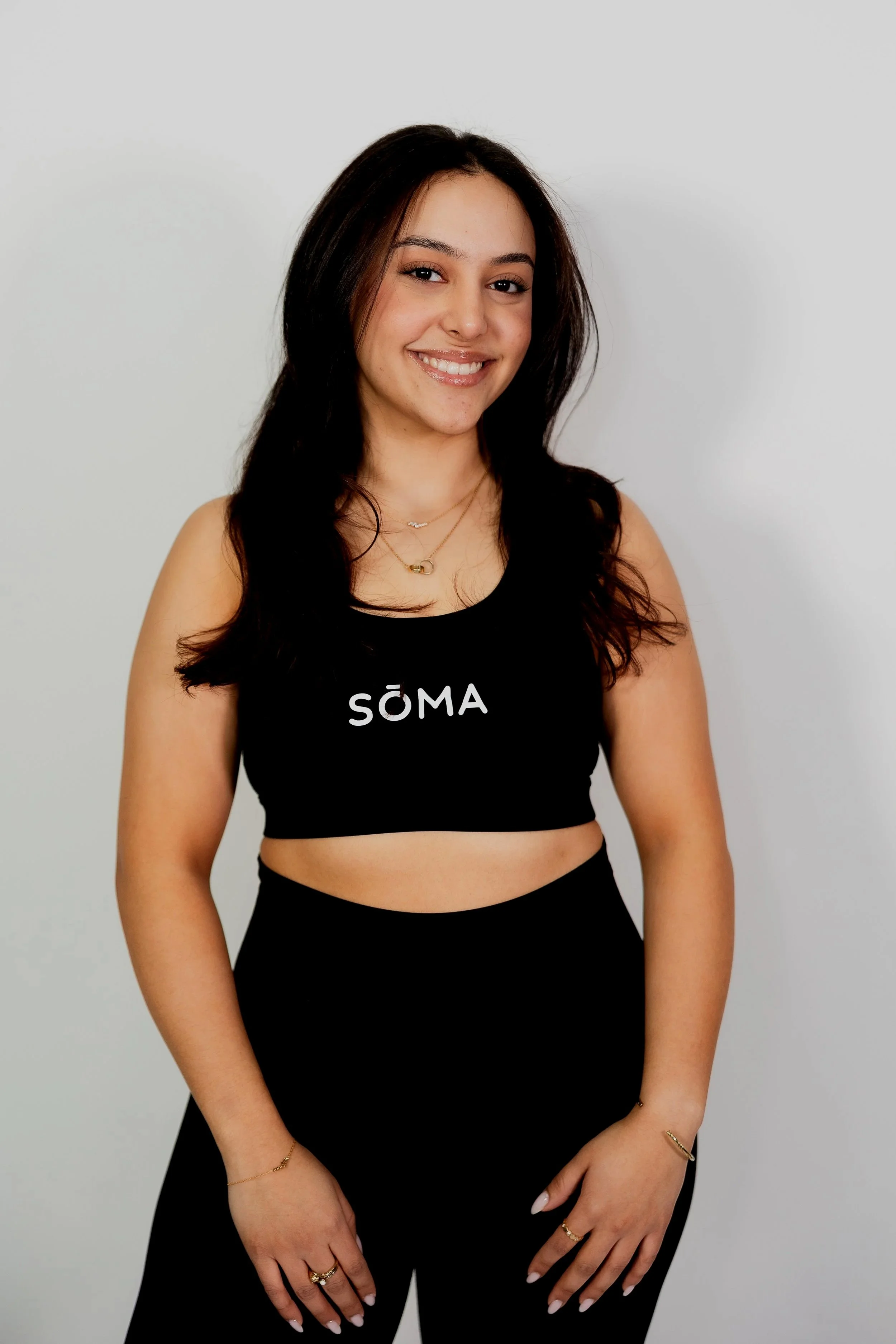 A young woman with long dark hair smiling, wearing a black crop top with 'SÖMA' written on it, gold jewelry, and black pants, standing against a plain white wall.