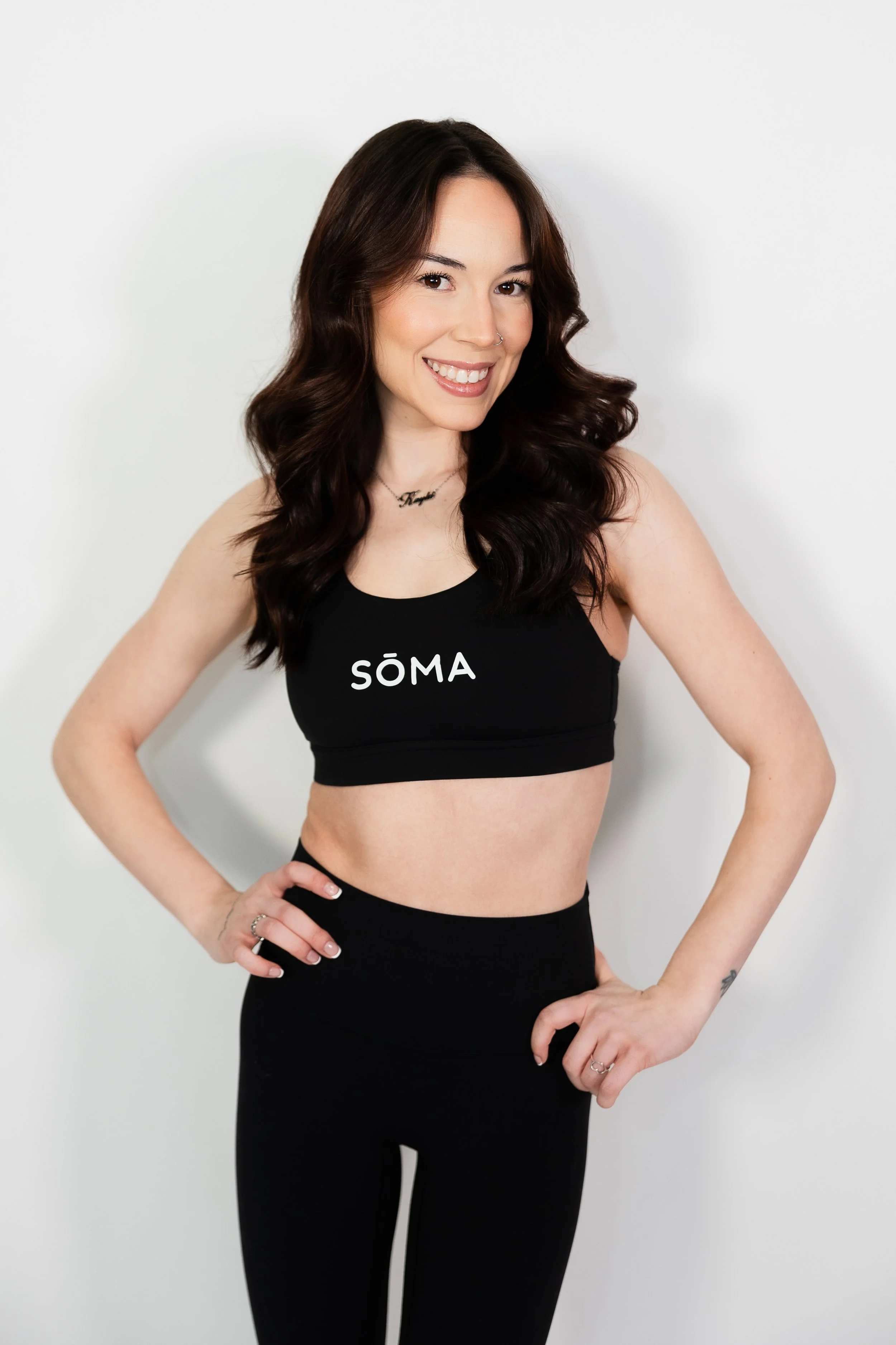 A woman with long dark brown hair wearing a black sports top with the word 'SÖMA' on it and black leggings, standing against a white wall, smiling and looking at the camera.