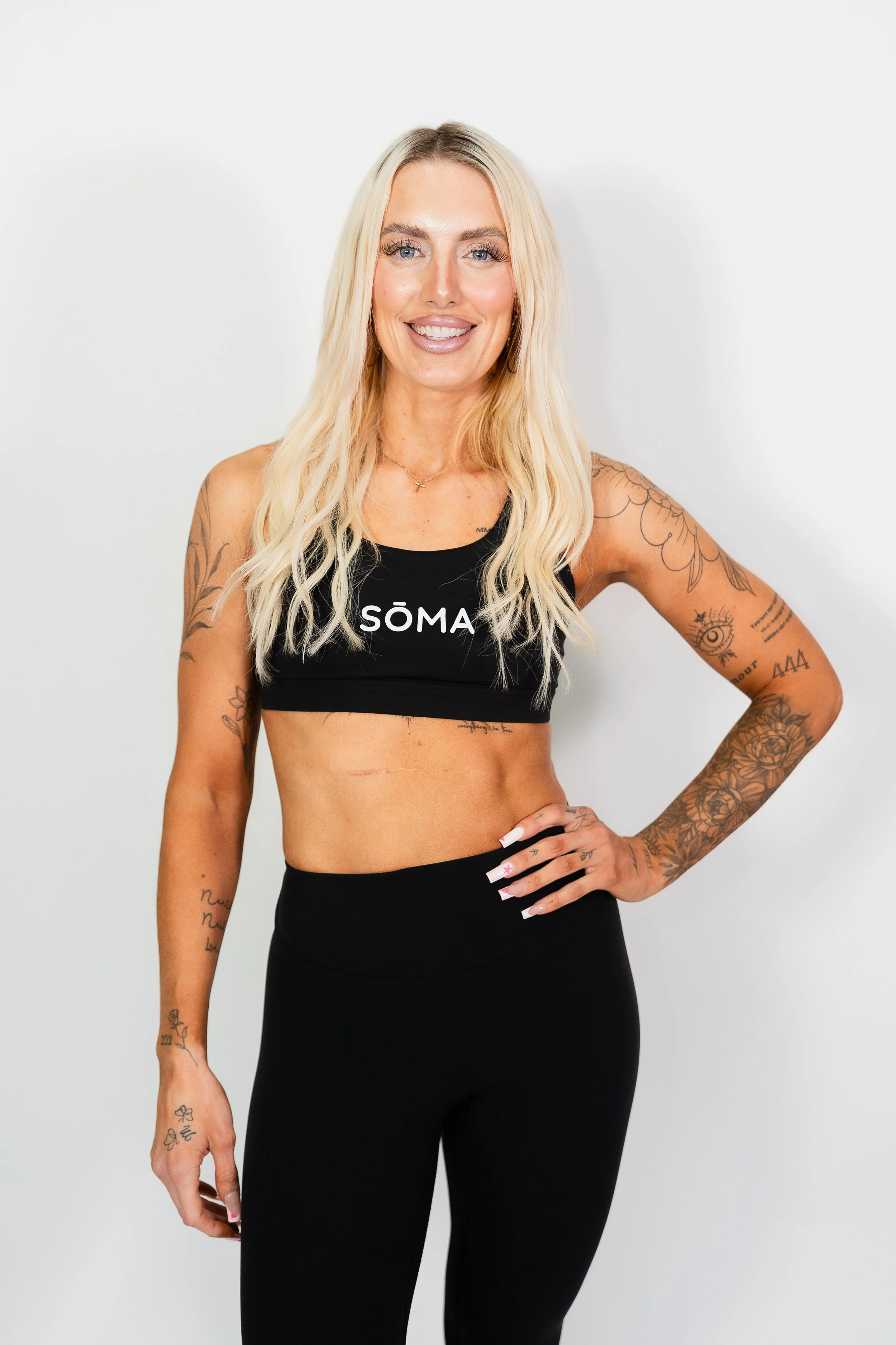 A woman with long blonde hair, blue eyes, and multiple tattoos on her arms, wearing a black workout top with the word 'SÖMA' on it, and black leggings, standing against a plain white background, smiling with her right hand on her hip.
