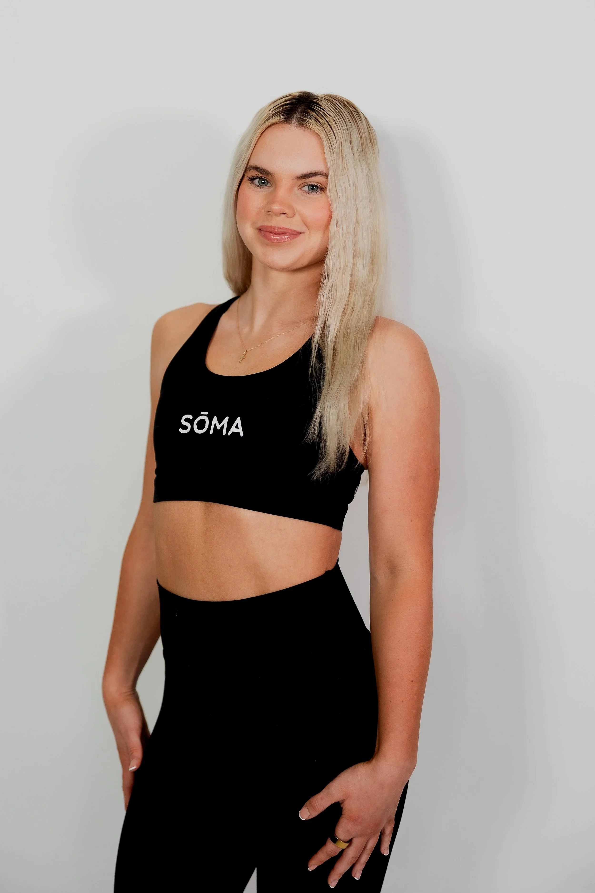 A young blonde woman wearing a black sports bra with the word 'SÖMA' written on it, standing against a plain white background.