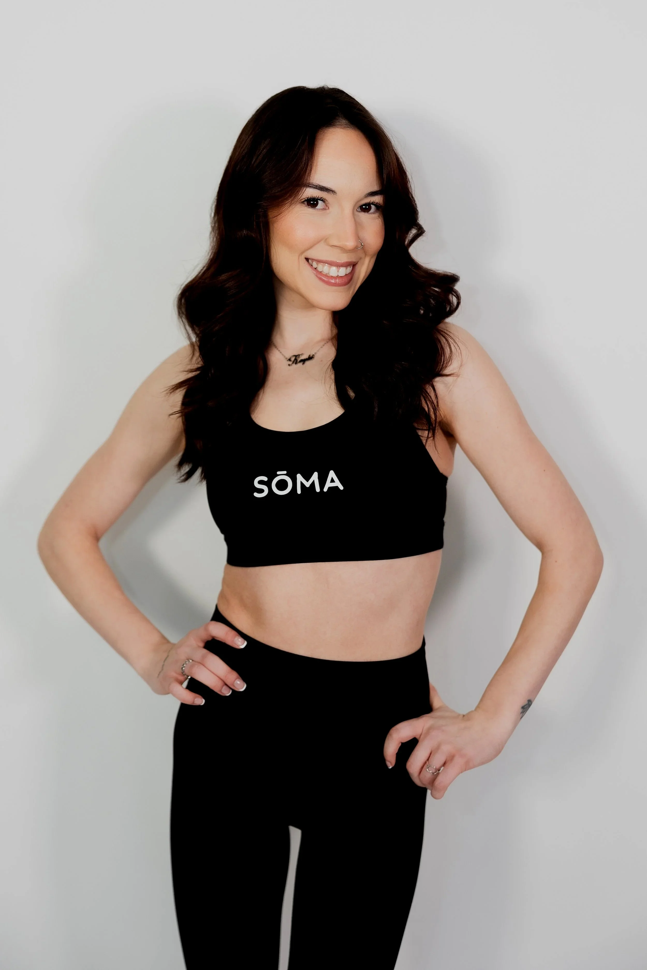 A woman with long wavy dark hair, smiling and wearing a black tank top with the word 'SÖMA' printed on it, standing against a plain white background.