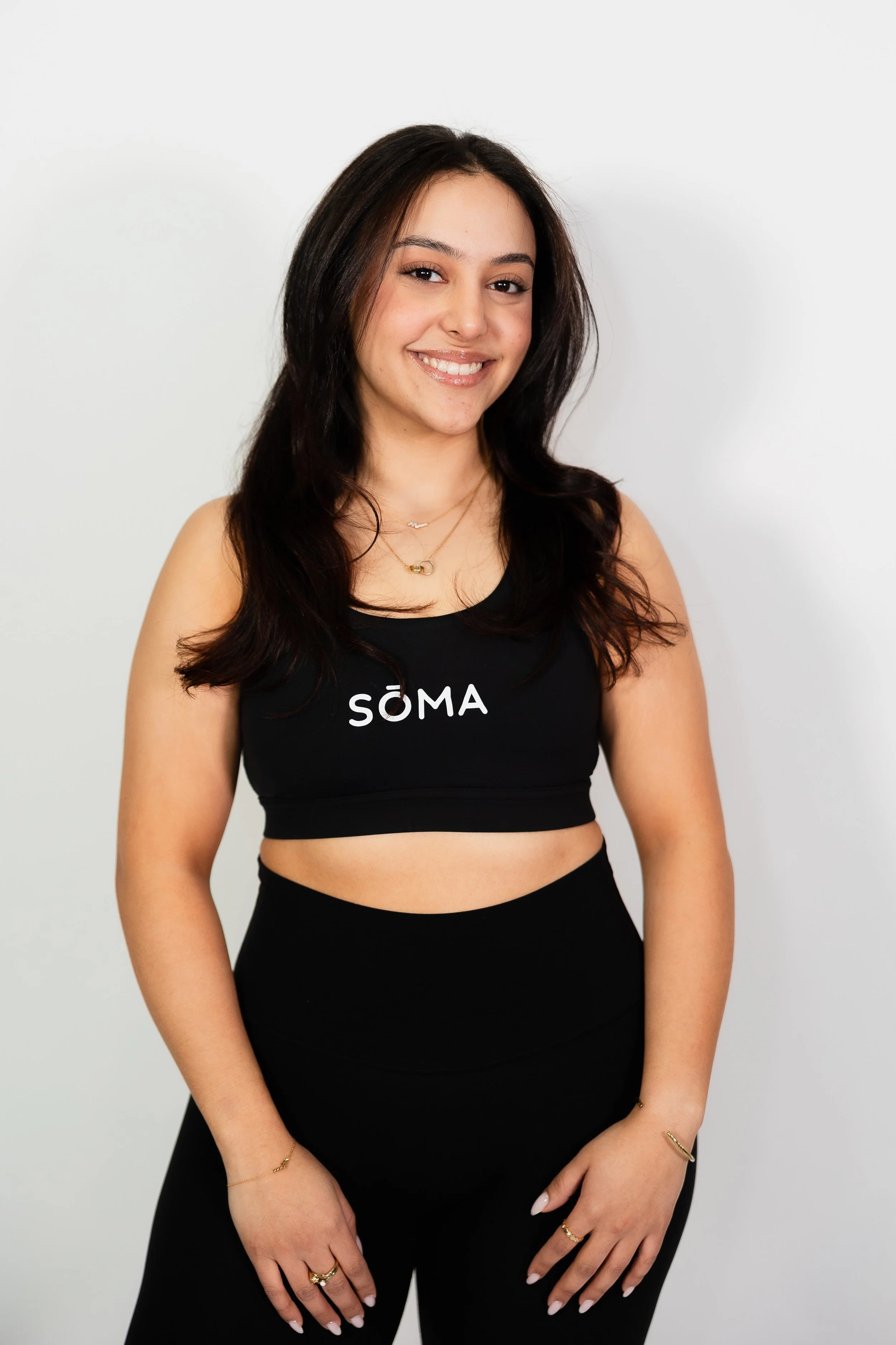 A young woman with long, dark hair and light skin is smiling and wearing a black sports bra with the brand name 'SÔMA' on it, paired with matching black leggings, standing against a plain white wall.