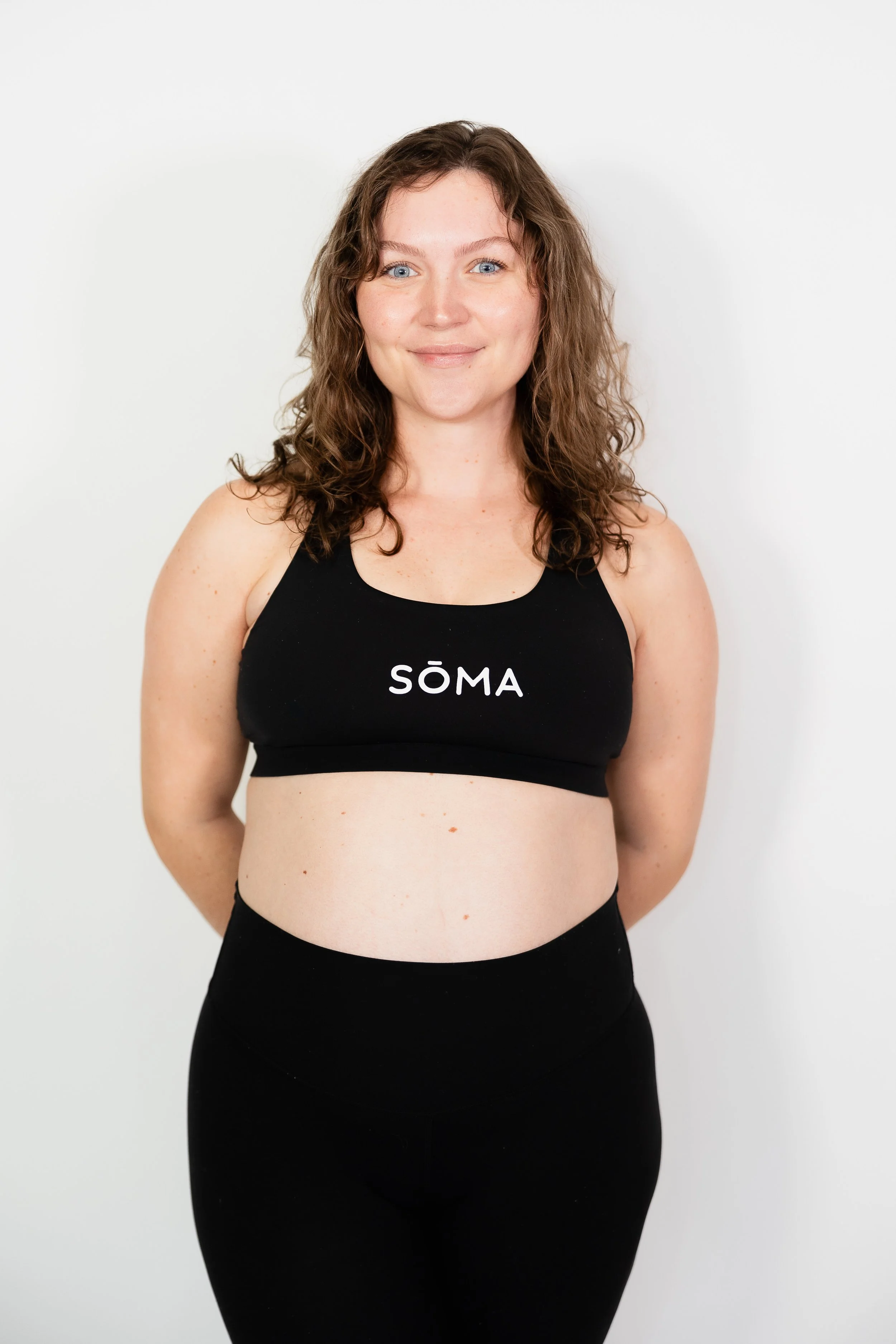 A woman with curly brown hair and blue eyes standing with her hands behind her back against a white background, wearing a black sports bra with the word 'SÖMA' on it and black high-waisted pants.