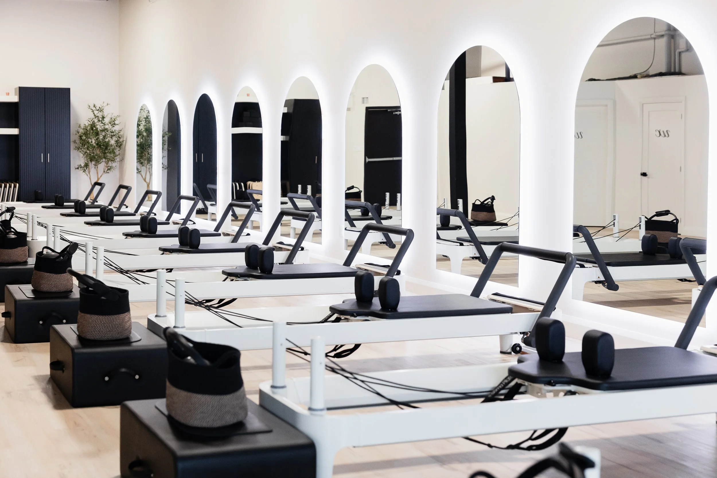 A modern Pilates studio with multiple reformer machines neatly arranged, white walls with arched mirrors, and potted plants, creating a clean and minimalistic fitness environment.
