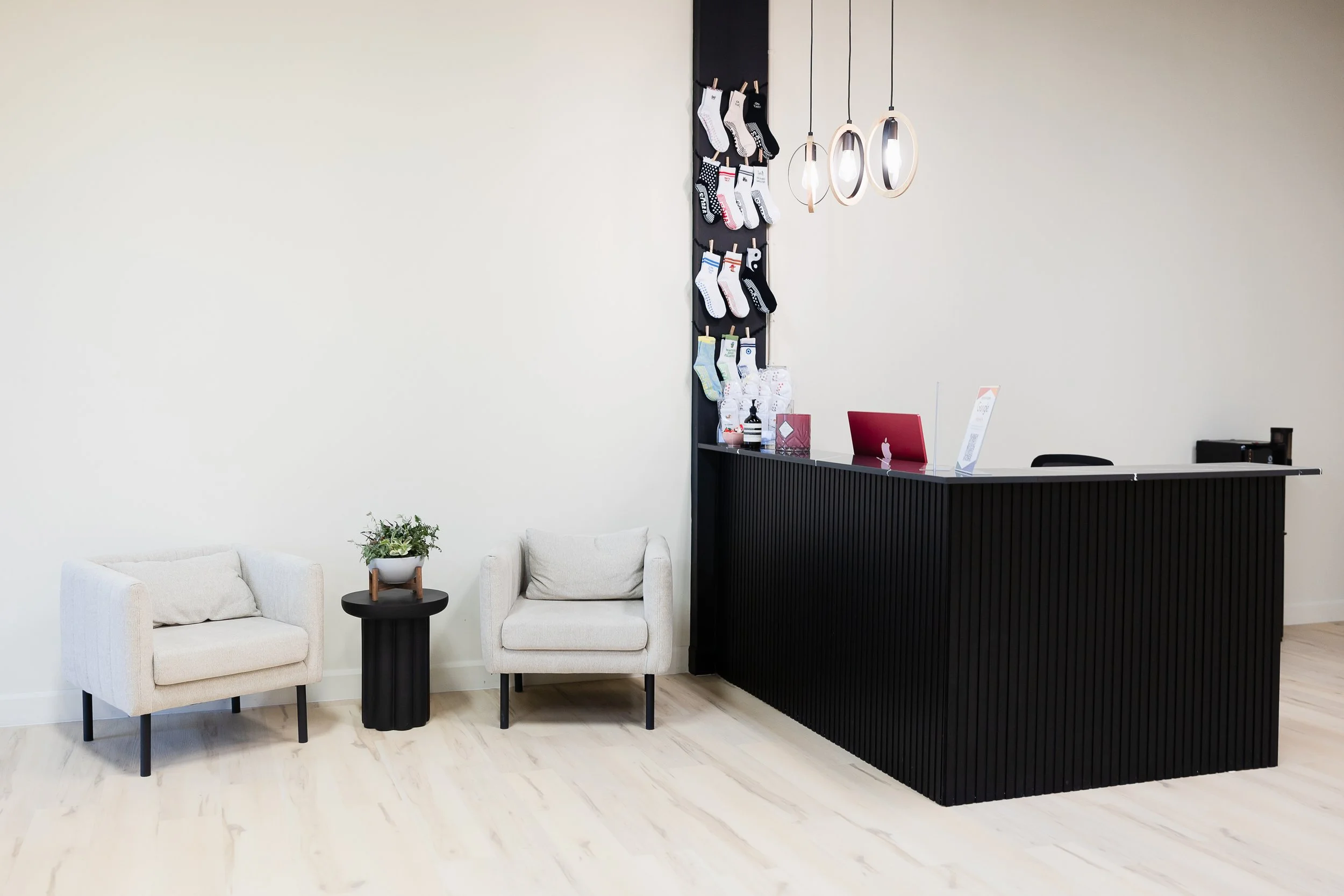 A modern office waiting area with two beige armchairs, a small round black side table with a potted plant, a black reception desk with a red laptop, and hanging pendant lights. Socks are displayed on a vertical rack behind the reception desk.