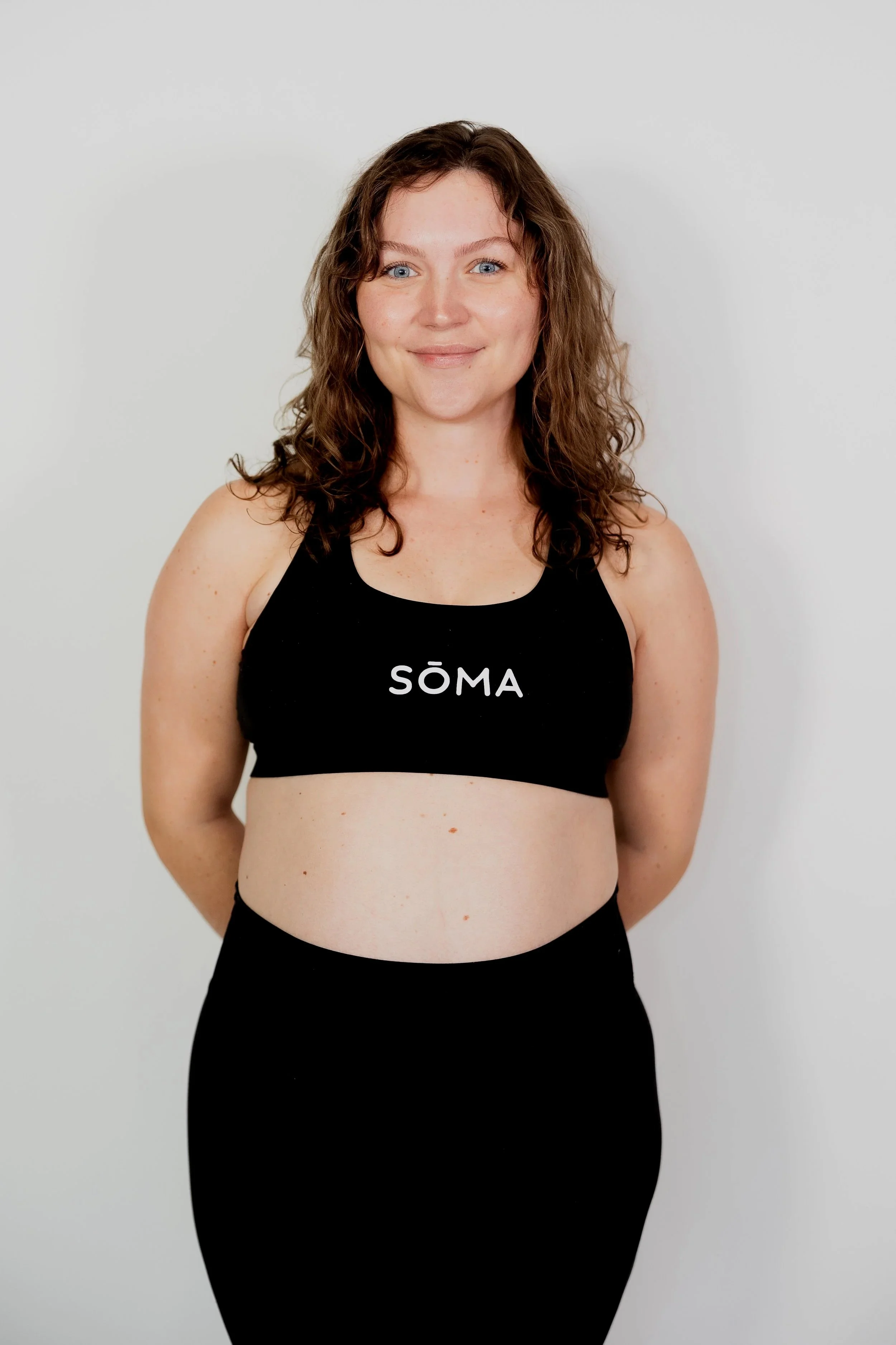 A woman standing against a plain light gray background, wearing a black sports bra with the word 'SÖMA' printed on it and black workout pants, smiling slightly at the camera.