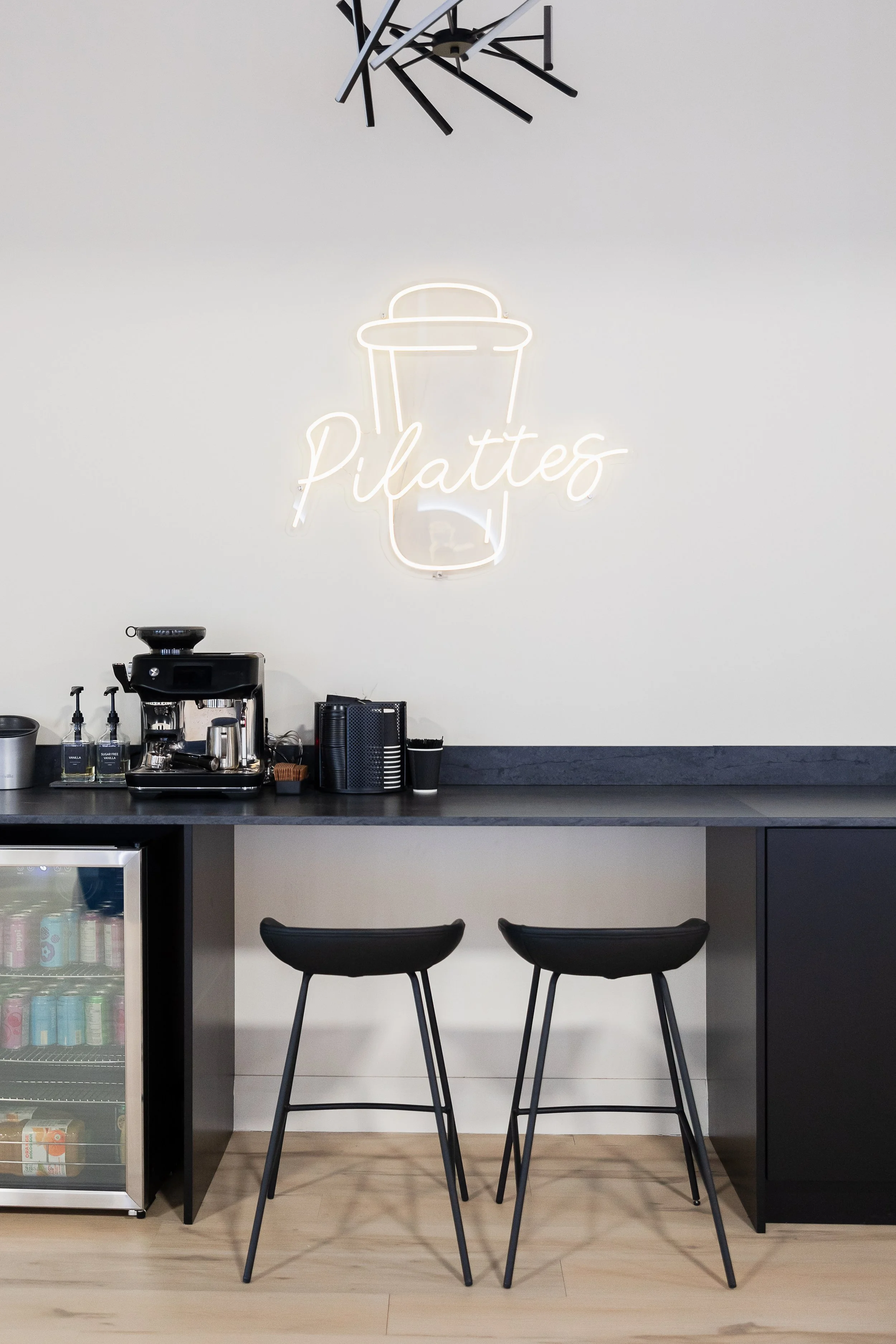 Neon sign on white wall displaying a coffee cup with the word 'Pilattes' in cursive. Coffee equipment on black countertop beneath the sign. Two black bar stools in front of the counter, and a small fridge with beverages on the left.