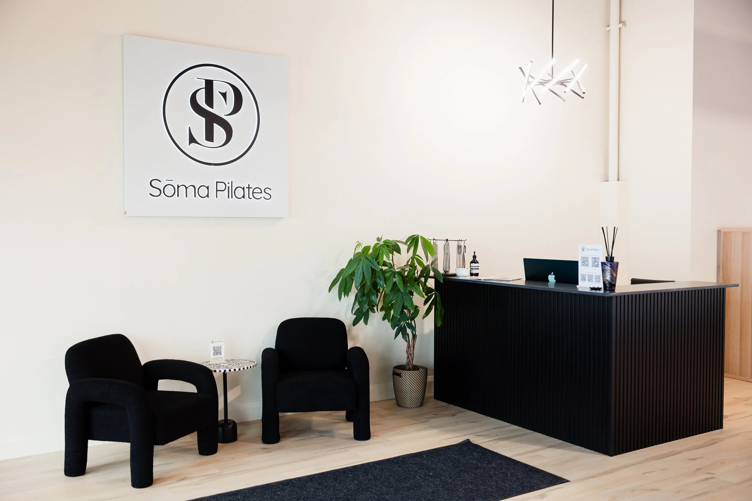 Modern lobby or reception area for Soma Pilates with black chairs, a small round table, green plants, a reception desk, and a large Soma Pilates sign on the wall.