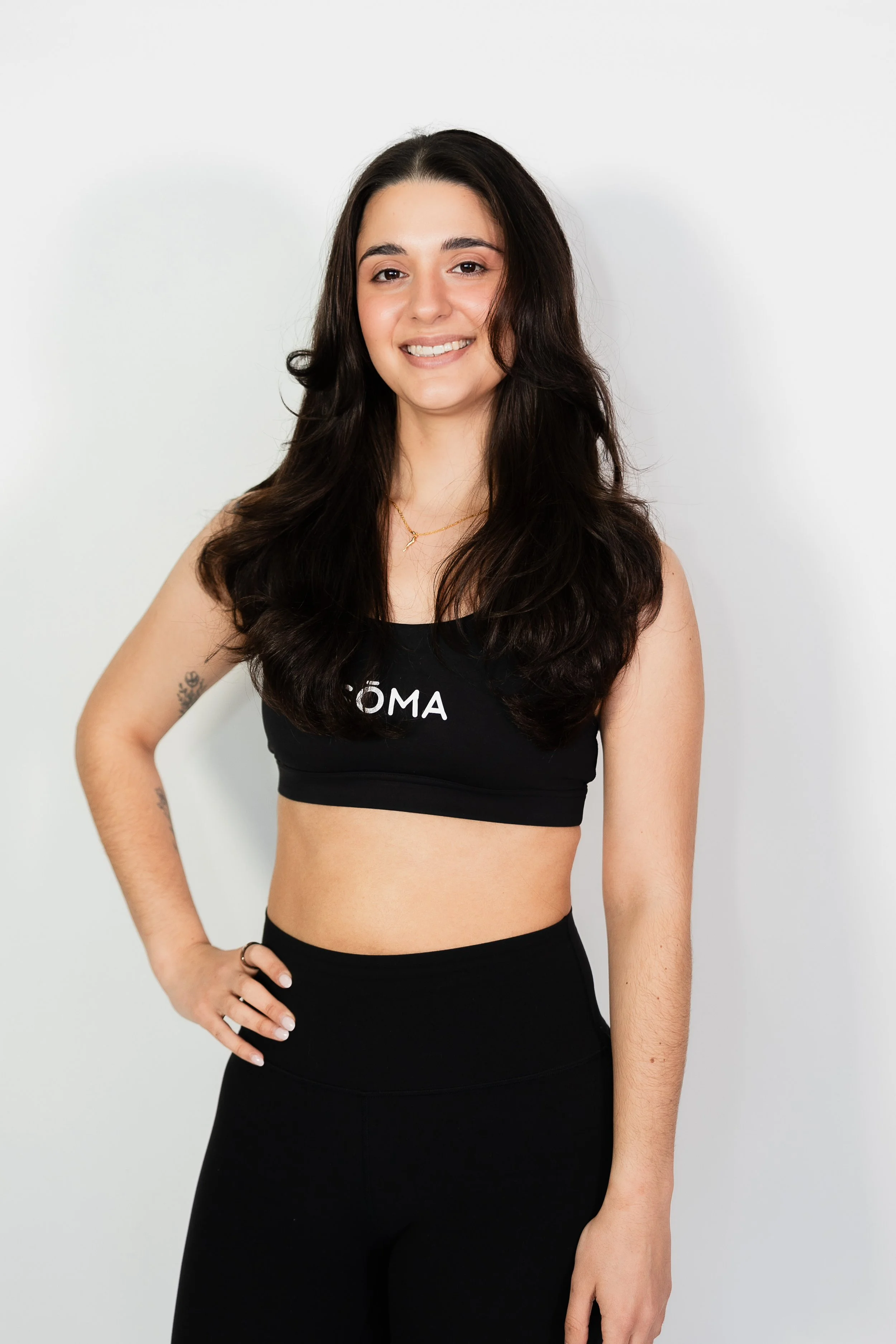 A young woman with long dark hair, wearing a black sports bra with white text and black leggings, smiling and posing with her left hand on her hip against a plain white background.