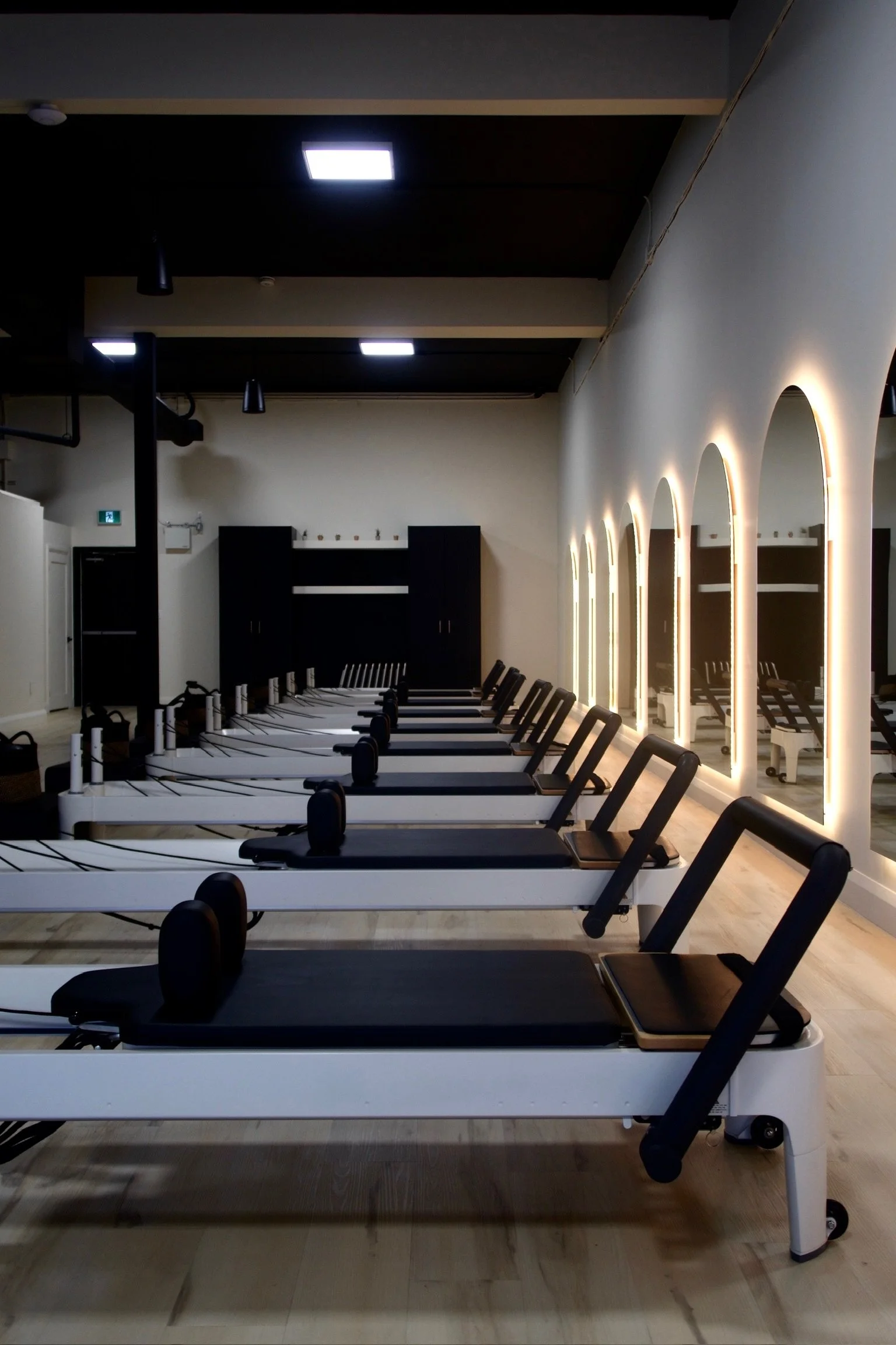 A pilates studio in ottawa with multiple Pilates reformer machines lined up, illuminated mirrors on the right wall, and ceiling lights.